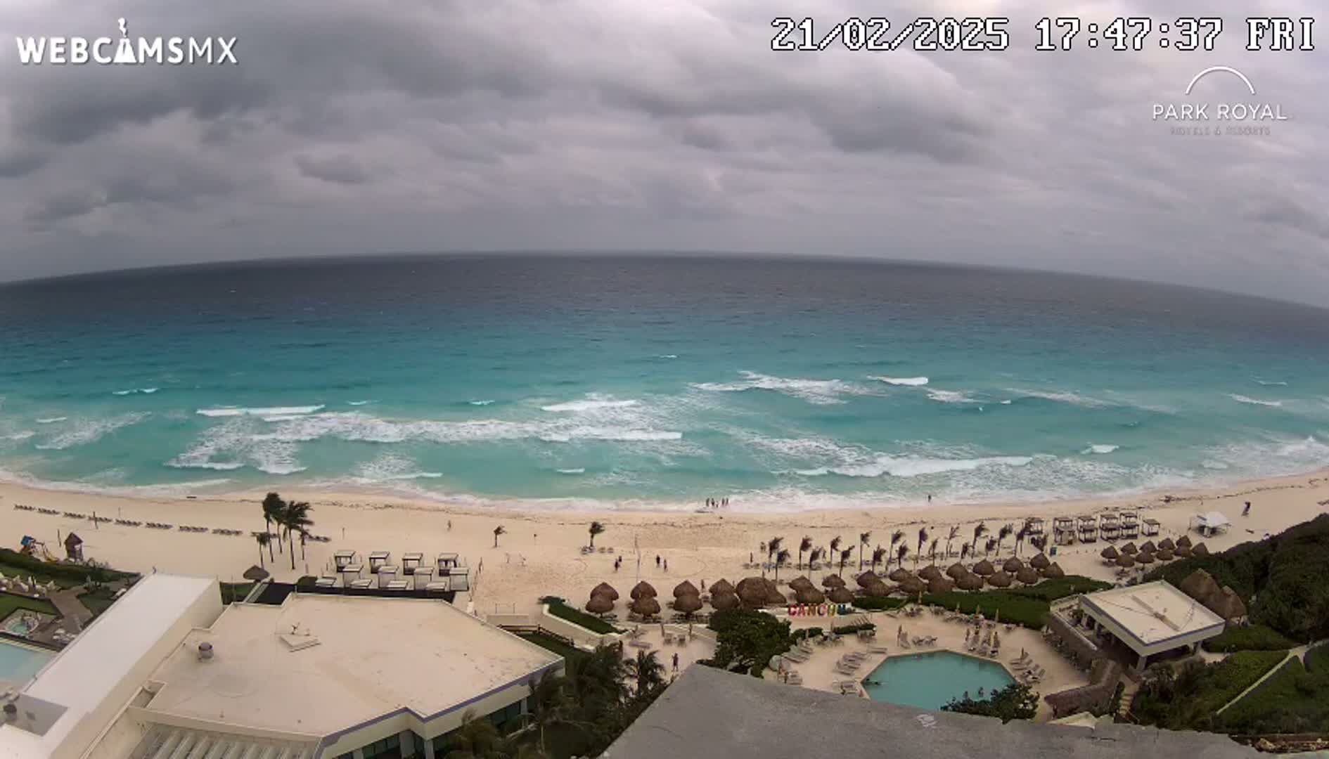 An ocean beach scene under an overcast sky shows a sandy beach with resort umbrellas and a swimming pool, all overlooking a turquoise ocean with small waves.