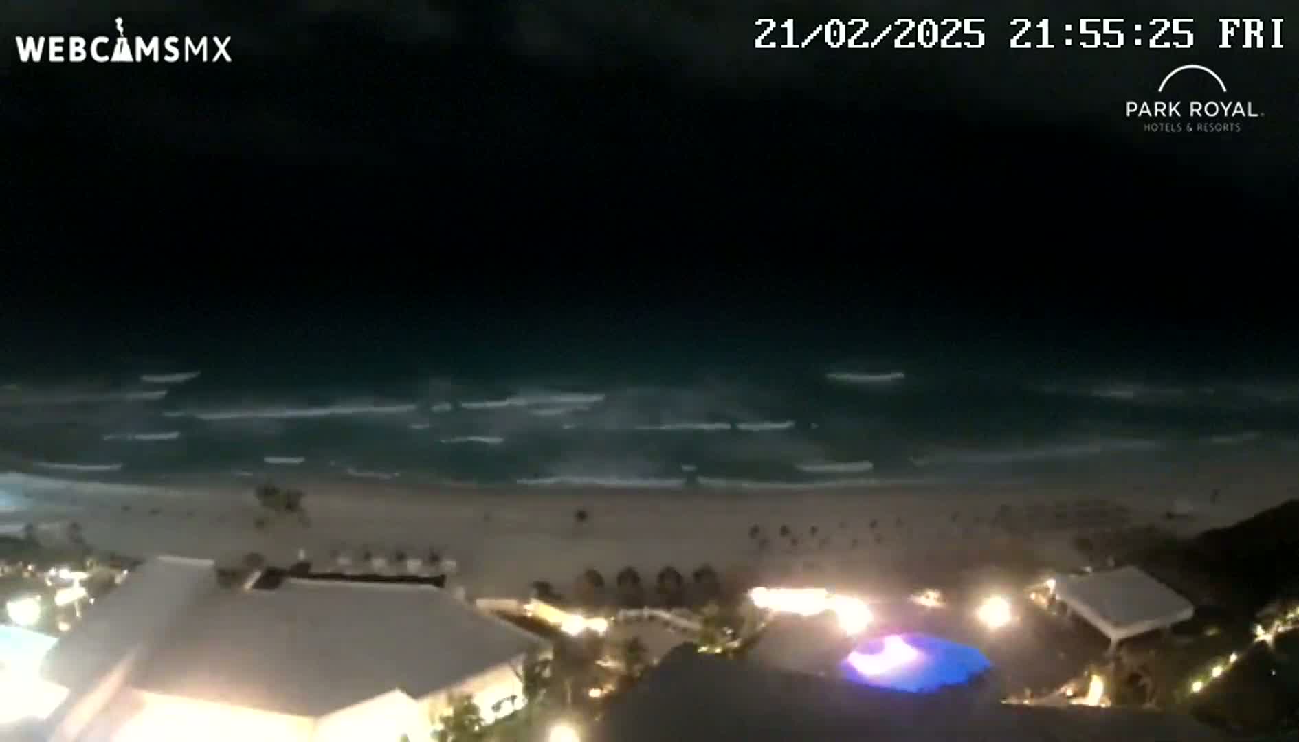 A nighttime view shows gently rolling waves on a beach, with illuminated buildings and a pool visible in the foreground.