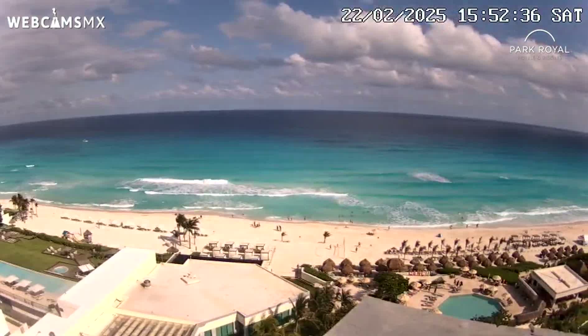 A partly cloudy day shows a beach with turquoise water, small waves, and a resort with a pool overlooking the ocean.
