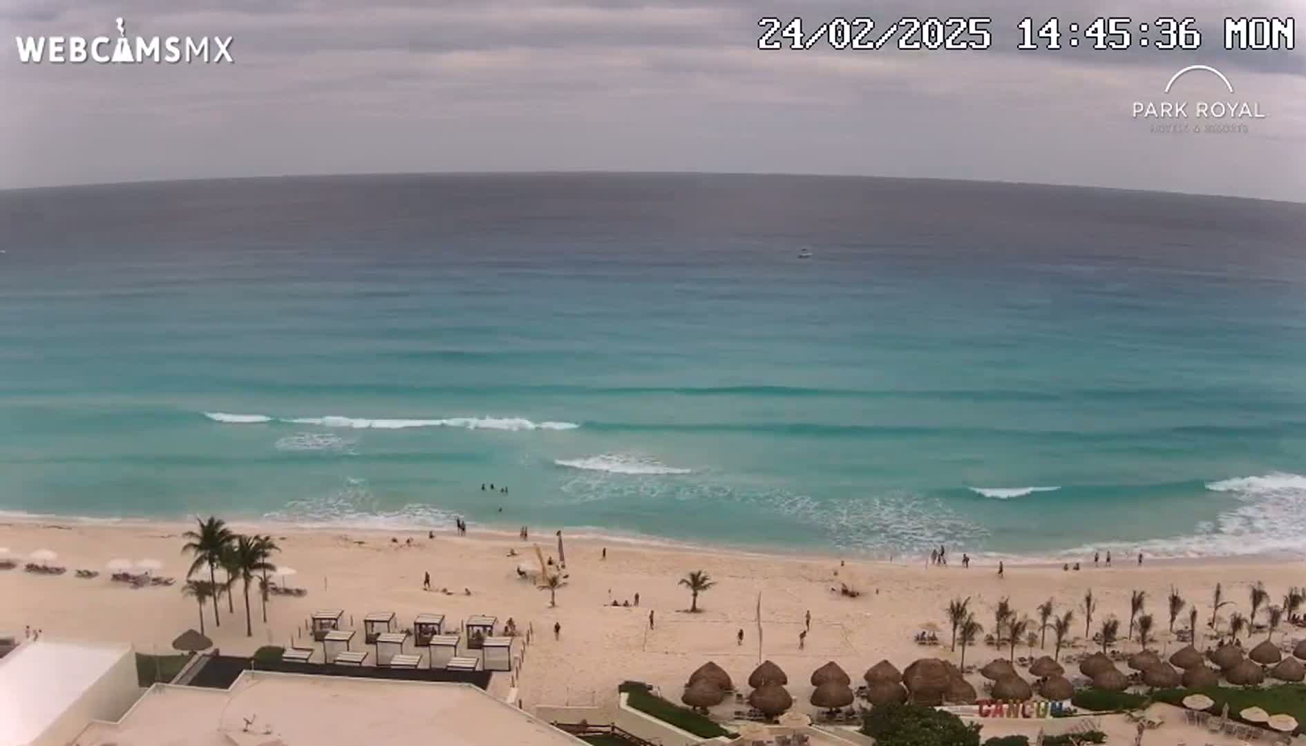 A beach with turquoise water and small waves under an overcast sky, viewed from a high vantage point, showing a sandy beach with some people and beach umbrellas.