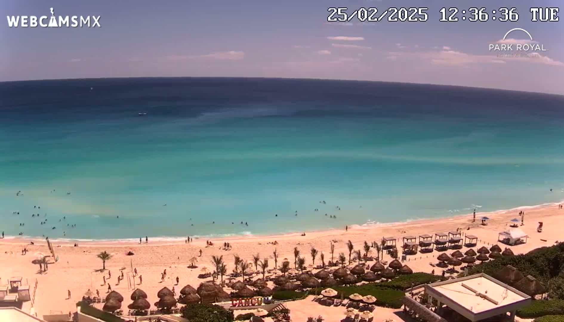A mostly sunny beach scene shows a tranquil turquoise ocean, a sandy shore with beach umbrellas and lounge chairs, and numerous people swimming and relaxing.