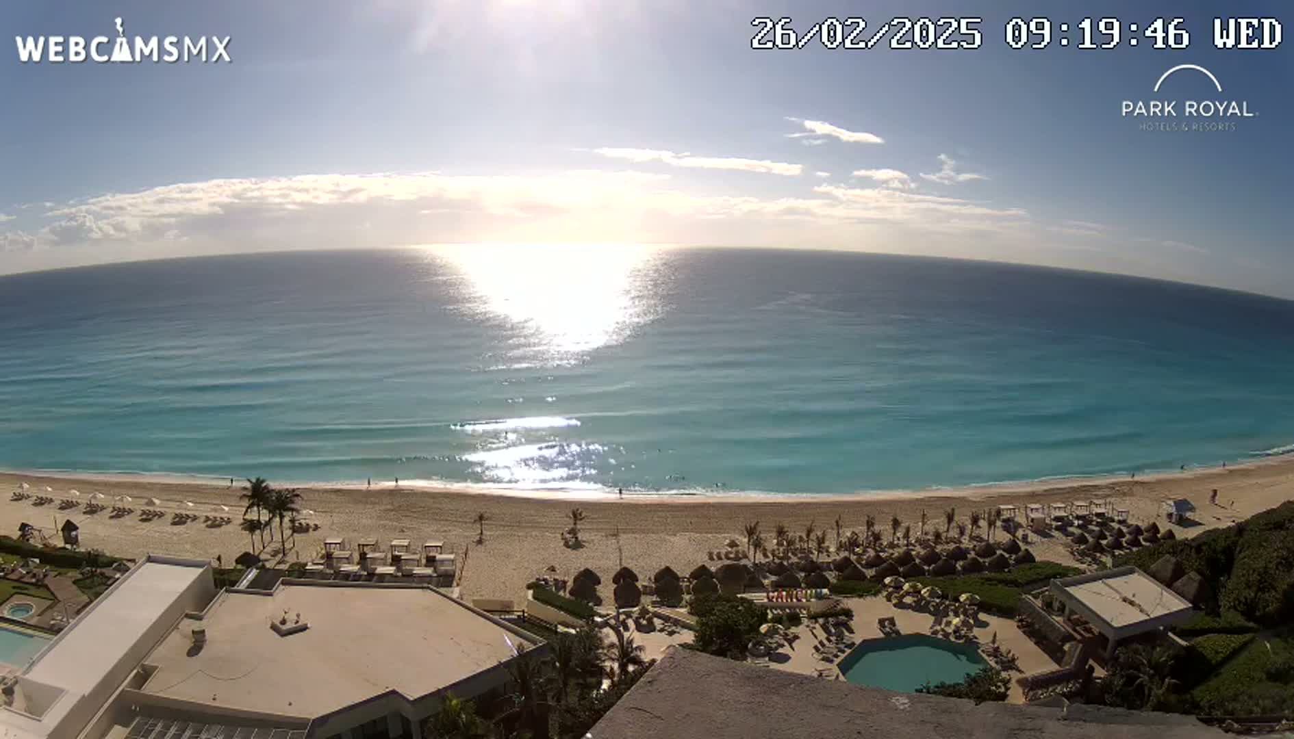 An aerial view shows a sunny beach with turquoise water under a mostly clear sky, overlooking a resort with a pool.