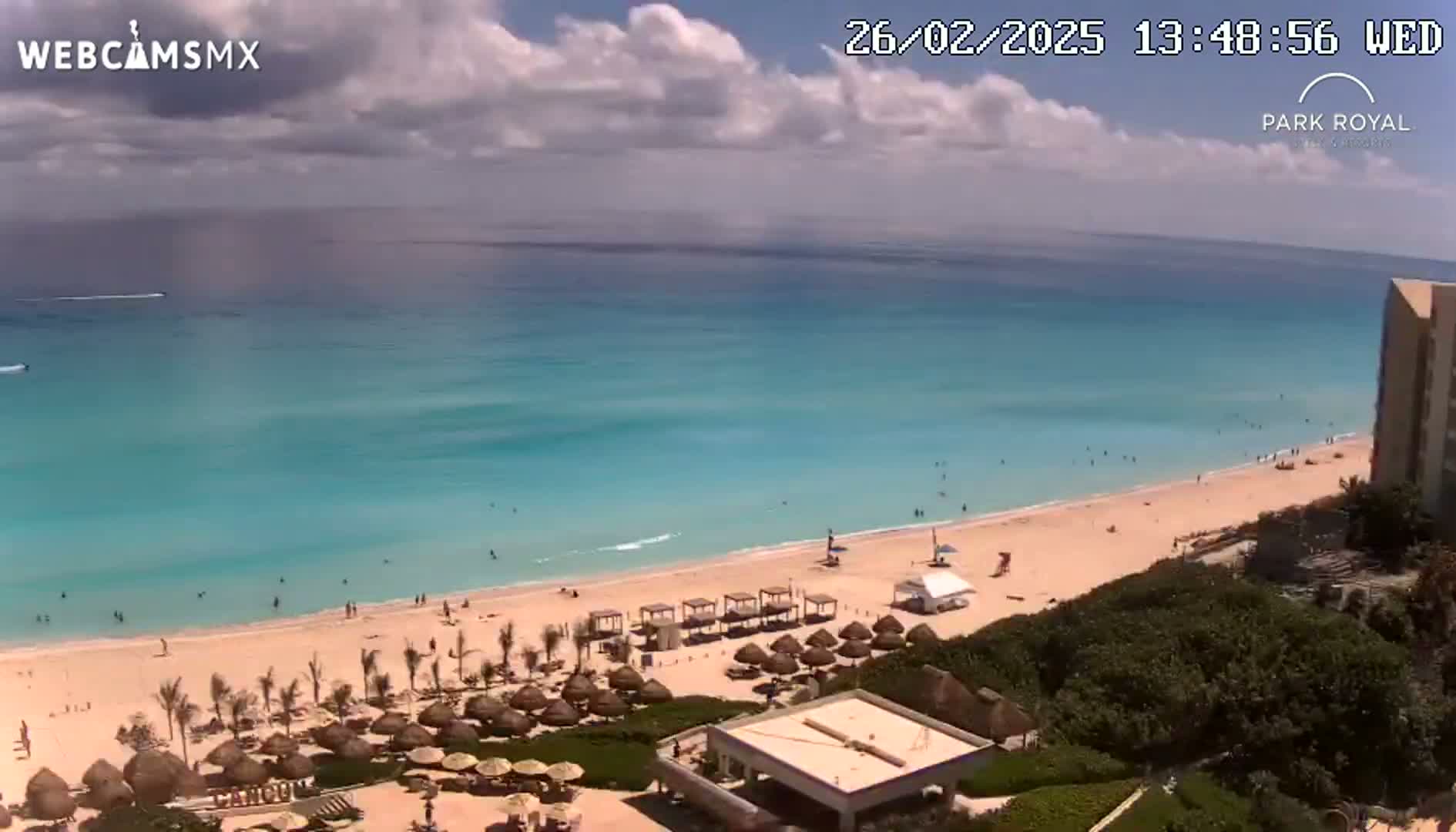 A partly cloudy day shows a pristine beach with turquoise water,  palm trees, beach umbrellas, and a few people enjoying the shore.