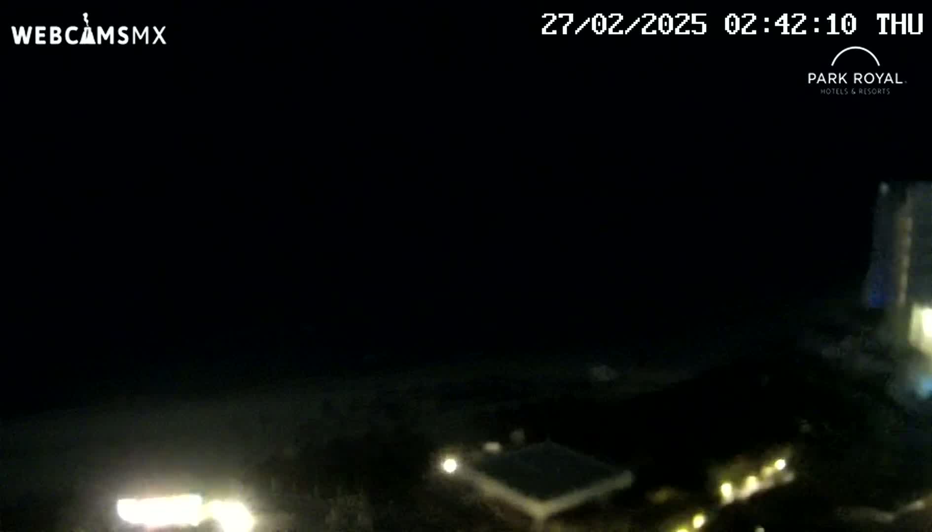 A nighttime, low-light view of a beach area with dimly lit buildings and a dark ocean under a clear, dark sky.