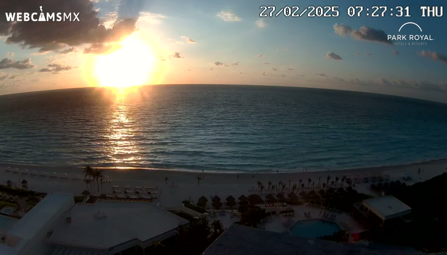 A sunrise over a calm ocean illuminates a sandy beach and a resort with a pool.