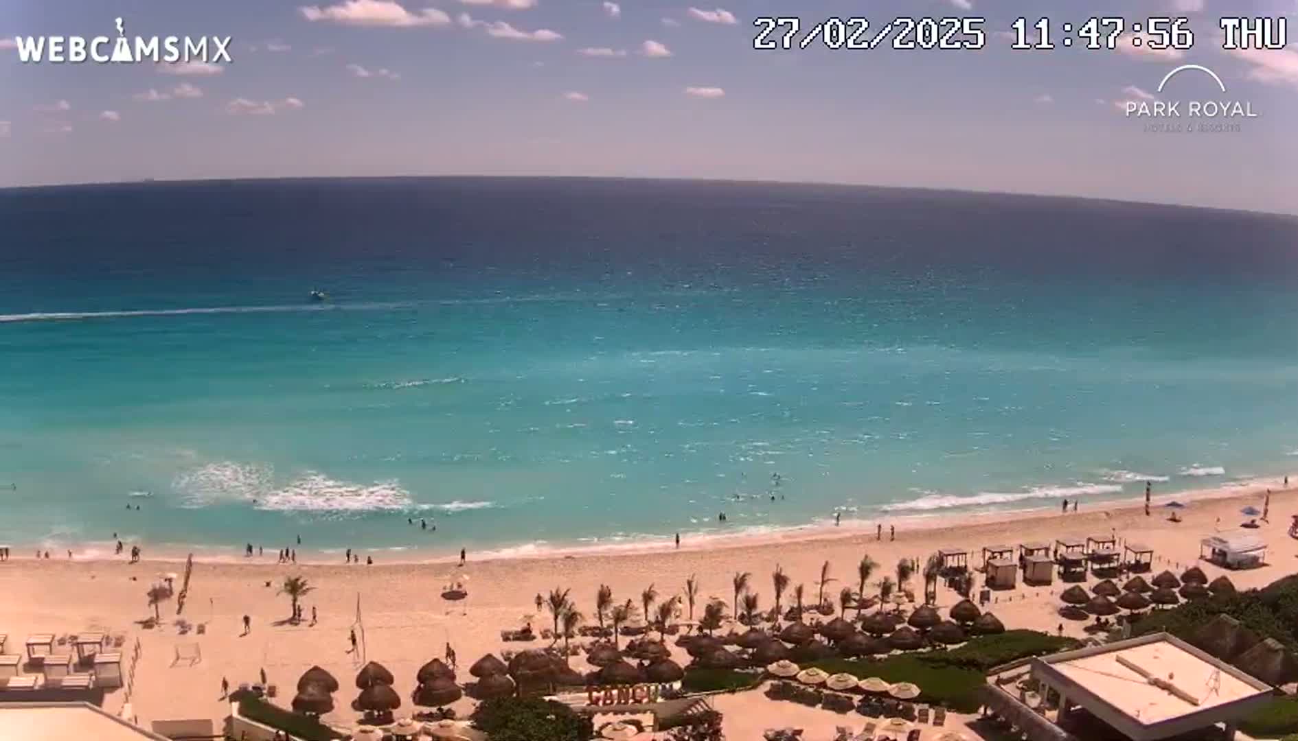 A sunny day at a beach resort shows a calm turquoise ocean lapping a sandy shore dotted with people and beach umbrellas.