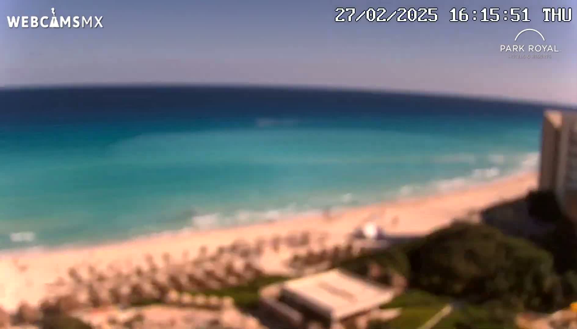 A beach with turquoise water under a clear sky, showing umbrellas and people on the sand and a building in the distance.