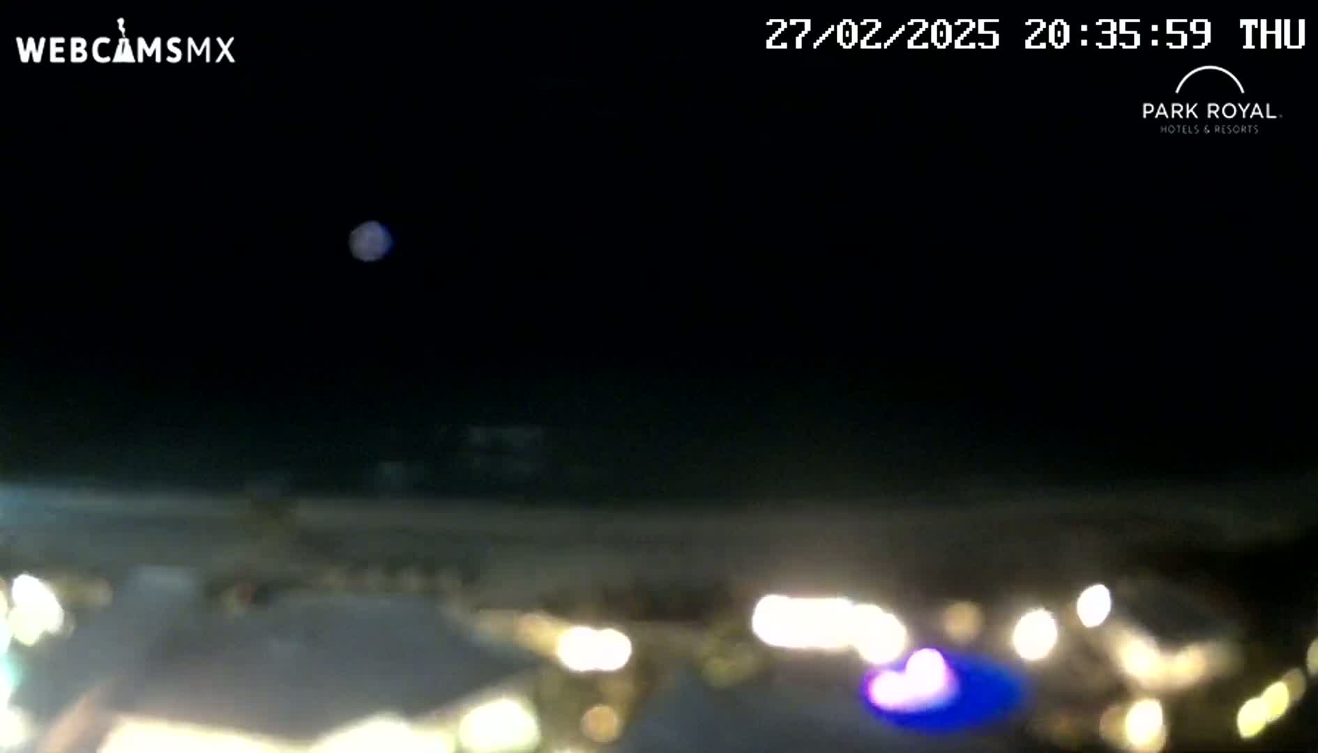 A nighttime, blurry view shows a dark, hazy sky above a beach with out-of-focus lights in the foreground.