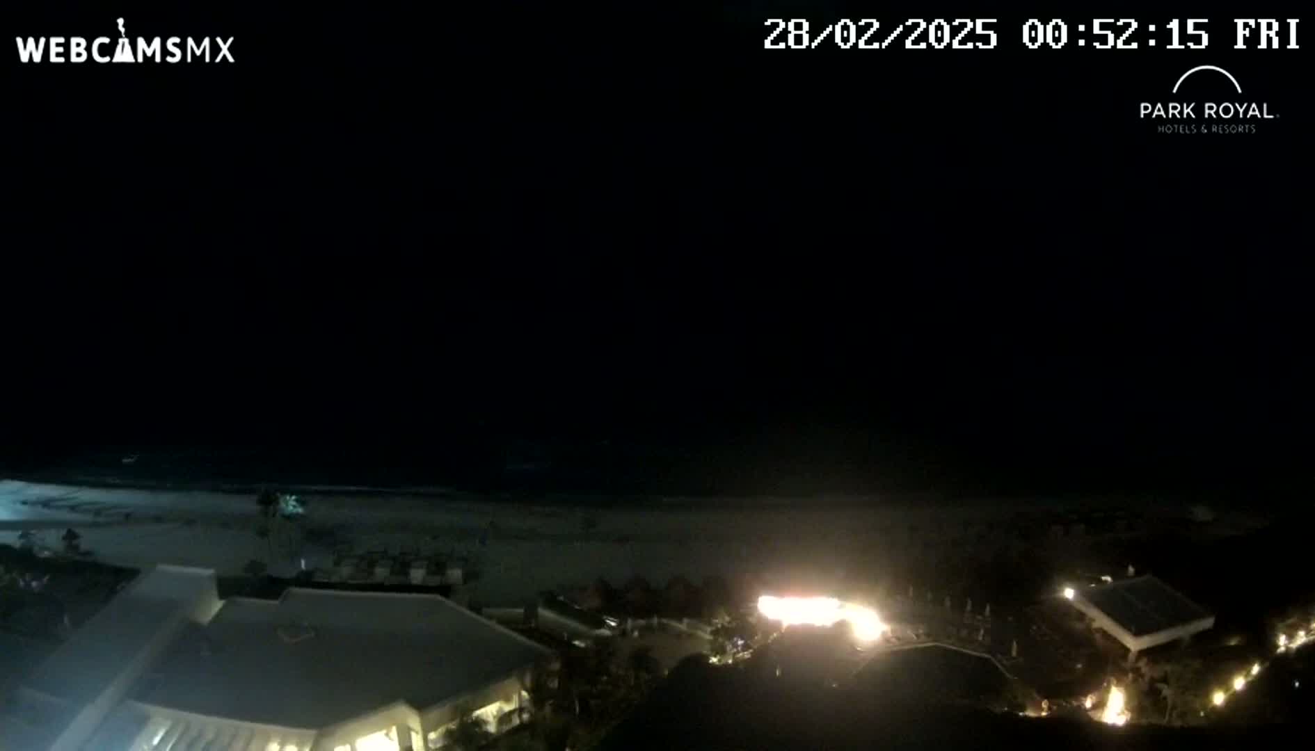 A nighttime view of a beach and resort, showing a dark ocean, a light-colored sandy beach, and lit buildings.