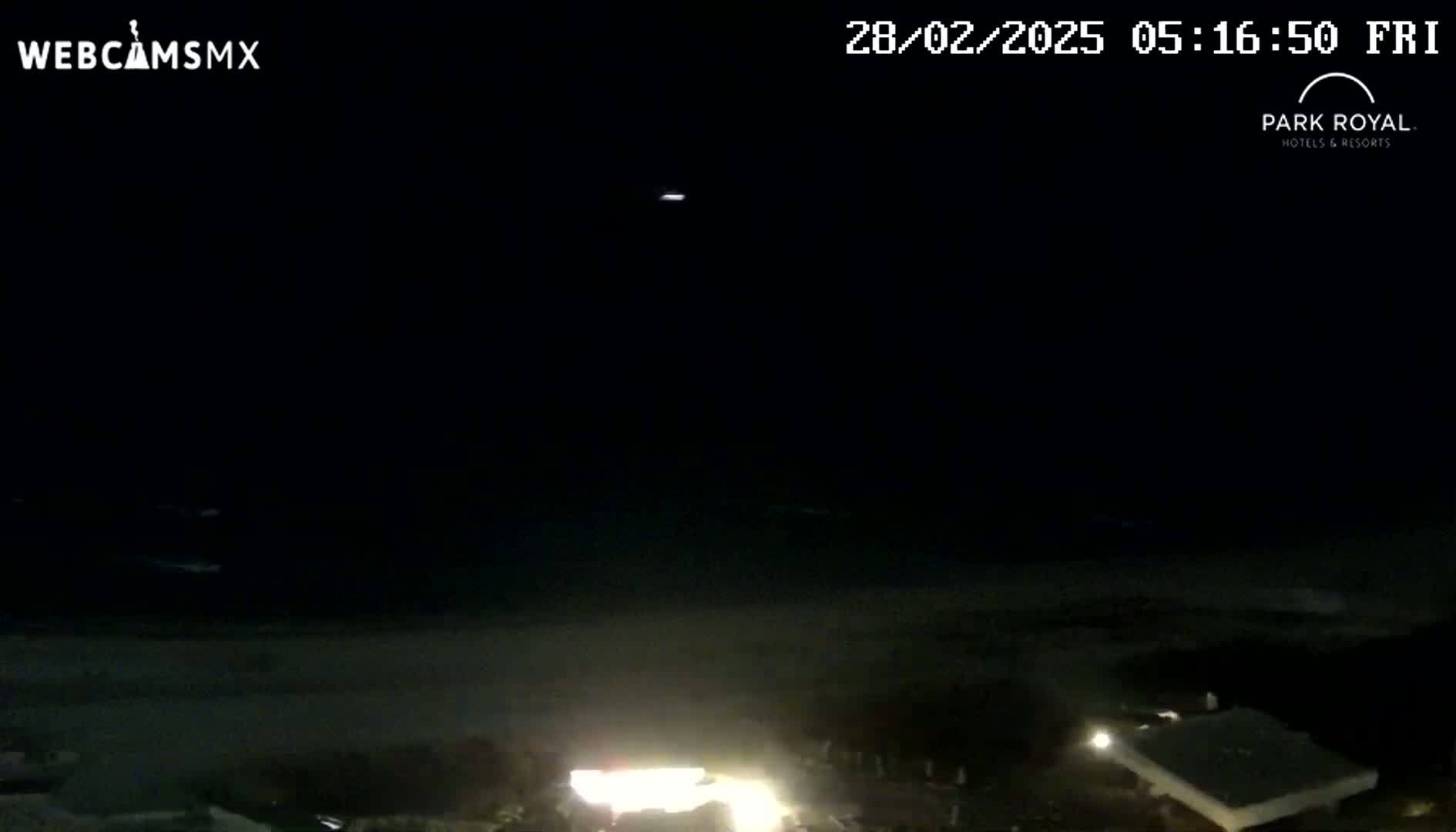 A nighttime view of a dark beach and ocean under a clear sky, with lights visible on the shore.