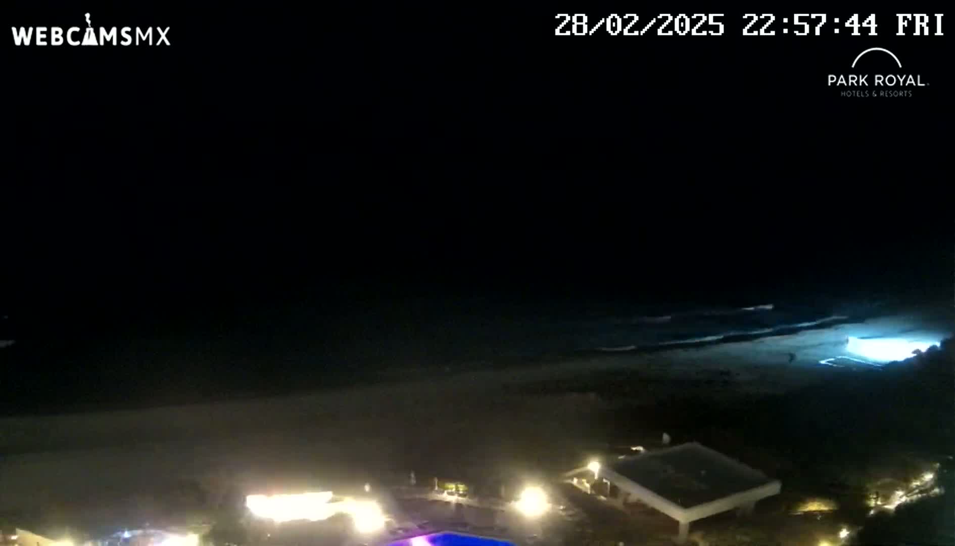 A nighttime coastal scene shows a dark beach with gently lapping waves under a clear, dark sky, with illuminated buildings and a swimming pool visible in the foreground.