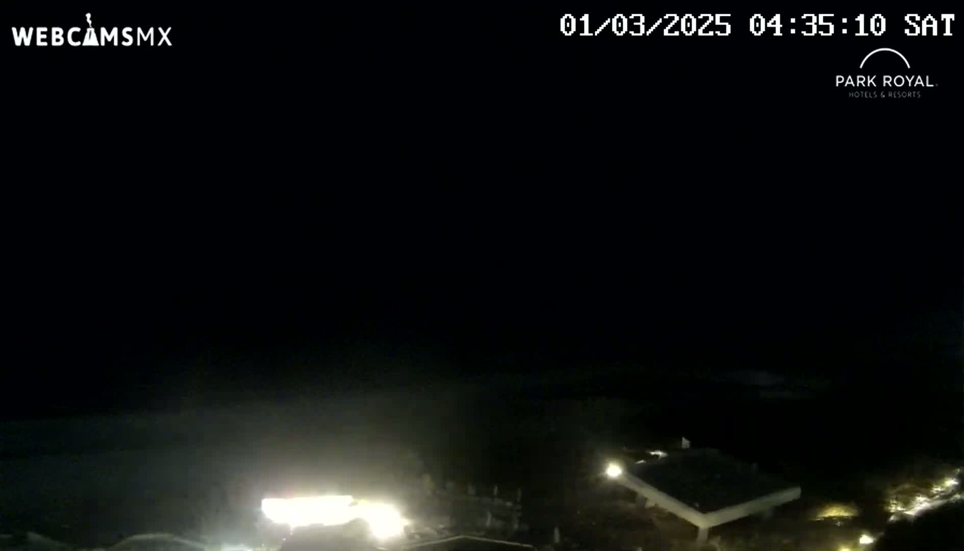 A nighttime, dark scene shows a blurry view of dimly lit buildings and a bright light source near the bottom, possibly from a vehicle, against a dark sky and ocean.
