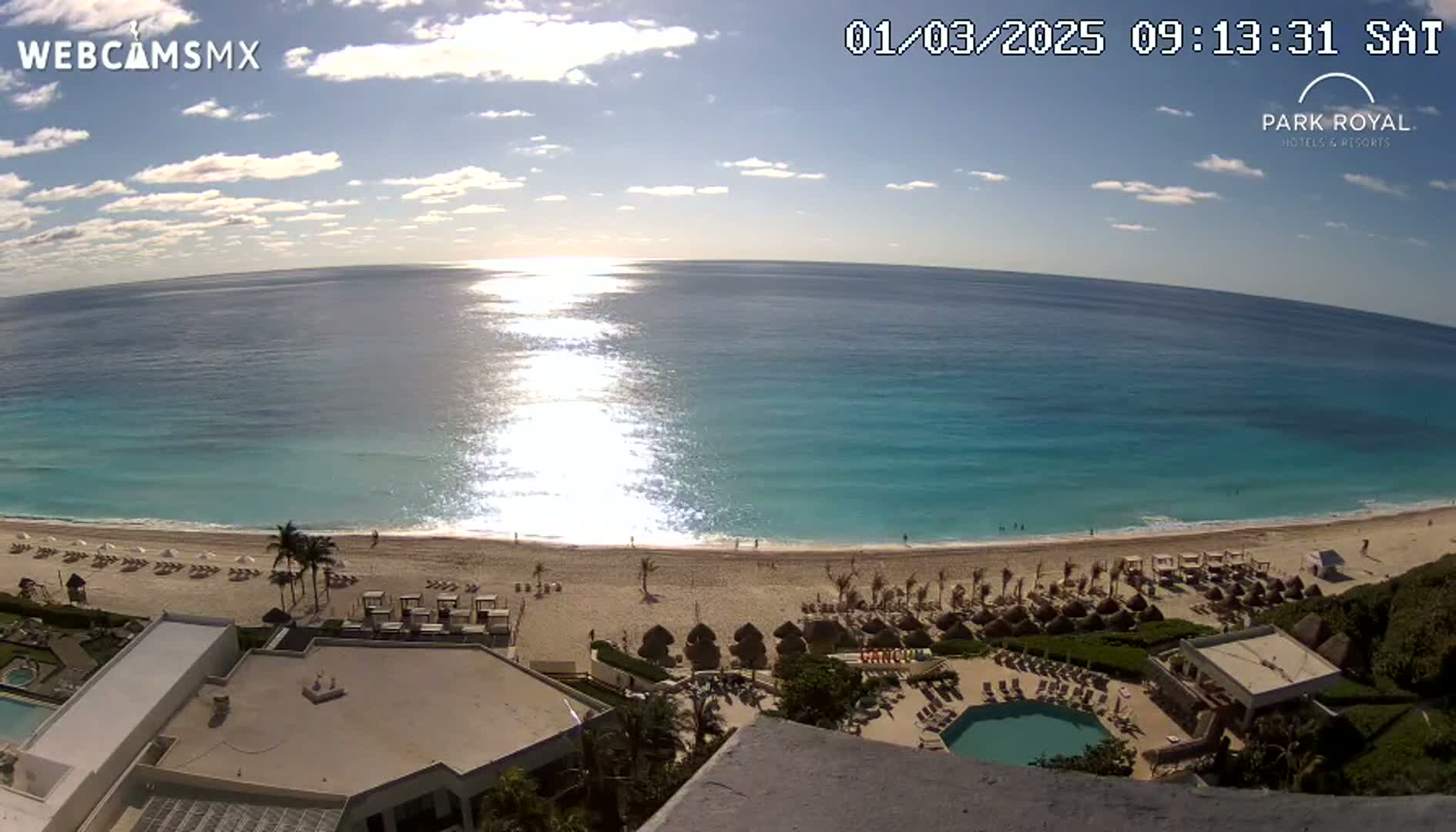 A sunny day shows a beach with turquoise water, a sandy shore lined with lounge chairs and umbrellas, and a resort with a swimming pool visible from an elevated viewpoint.