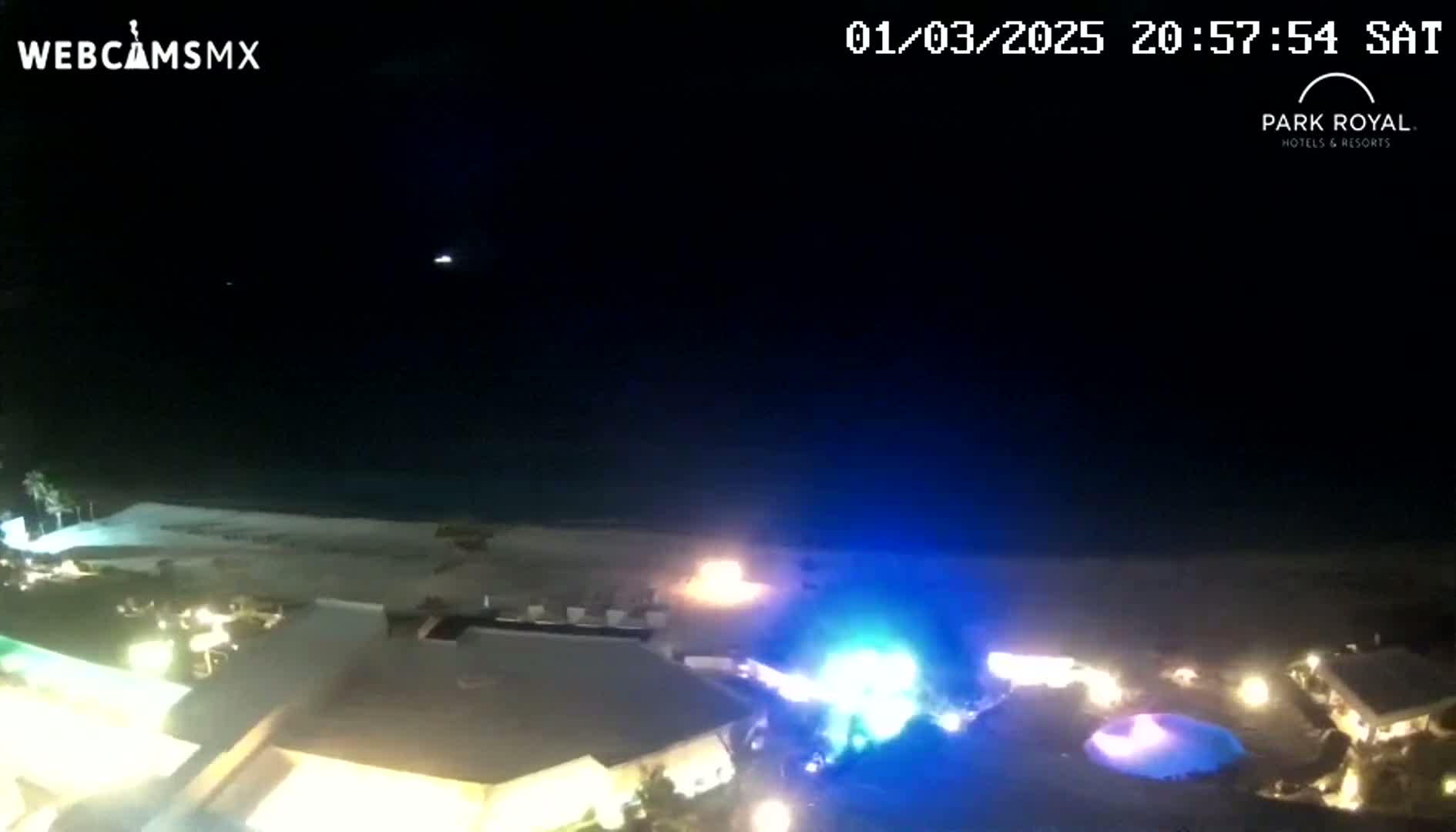 A nighttime aerial view shows a beach, resort buildings with lights on, and a bonfire or fireworks display on the sand, under a clear, dark sky.