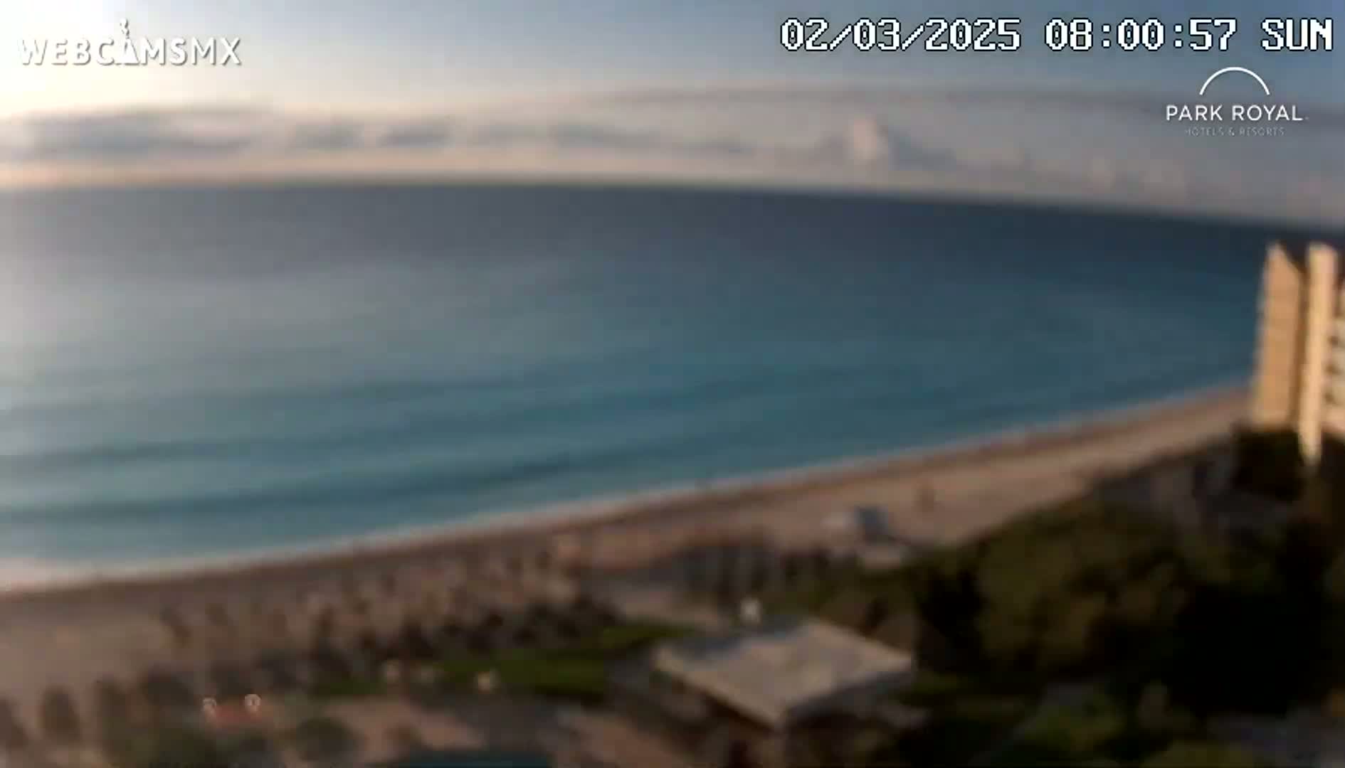 A blurry, high-angle view shows a mostly sunny day at a beach with calm ocean waves, a sandy shoreline, and some vegetation near a building.