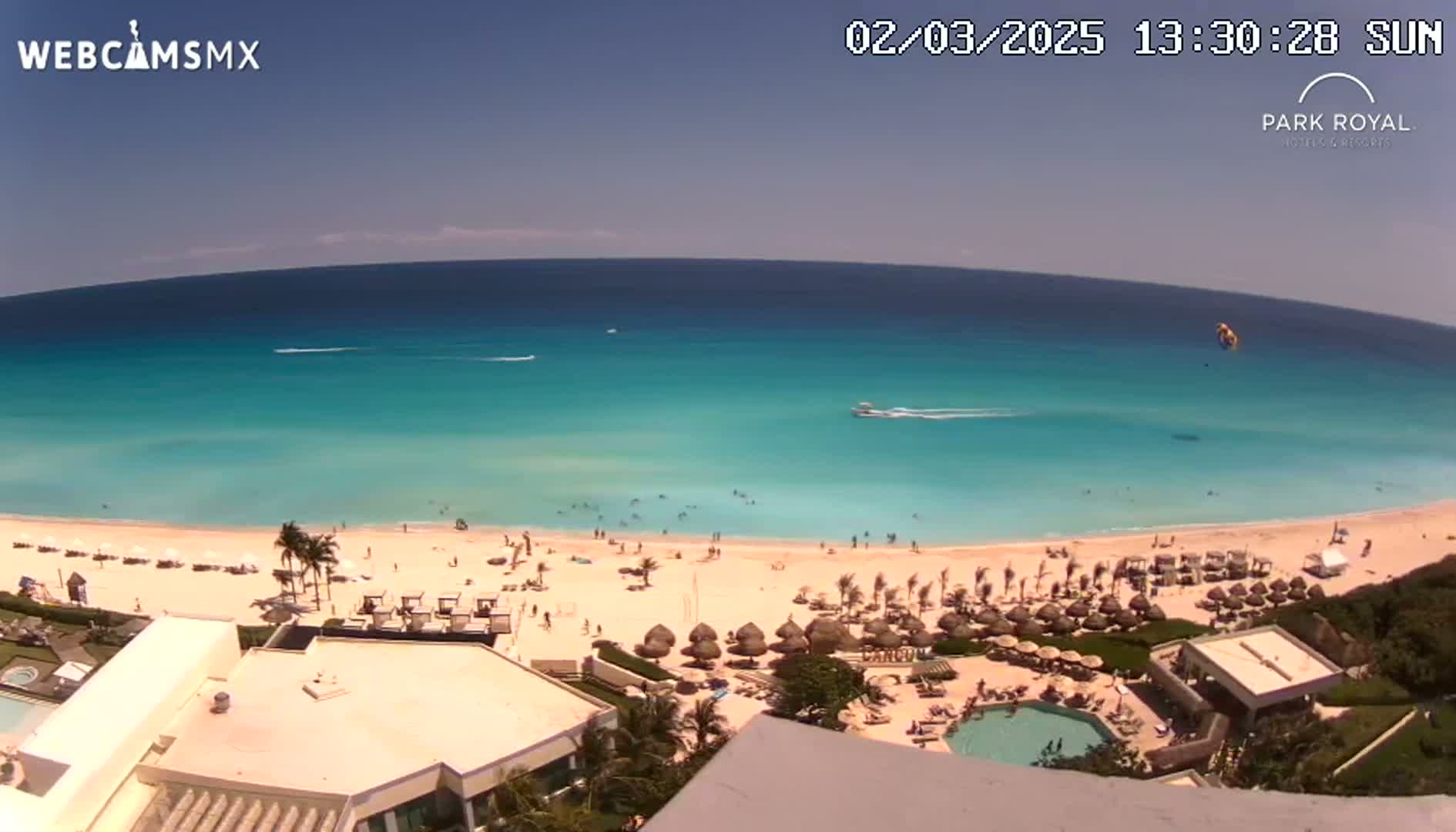 An aerial view shows a beach with turquoise water, a sandy shoreline dotted with people and beach umbrellas, and a resort with a pool under a sunny sky.