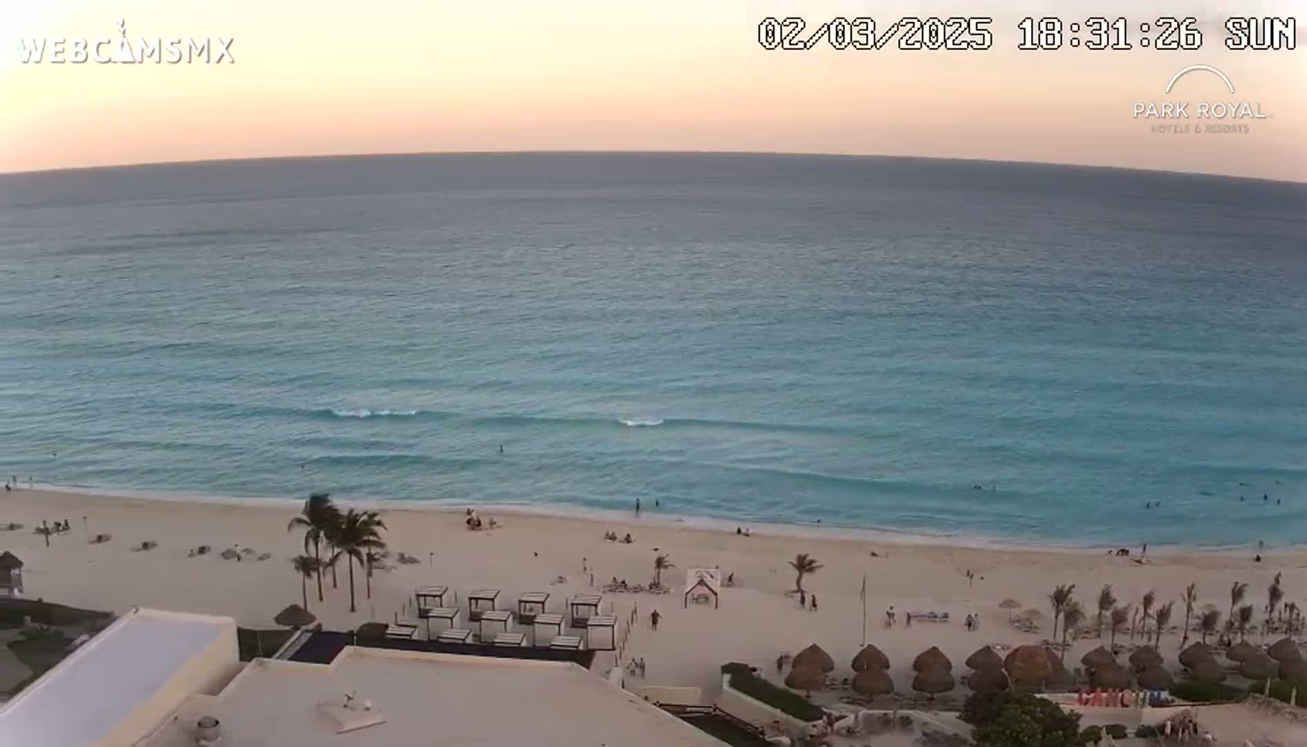 A beach with calm turquoise water under a sunset sky, showing palm trees, beach umbrellas, and a few people on the sand.