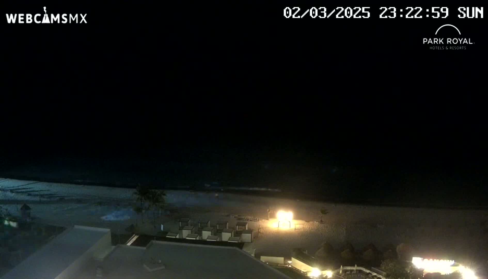 A nighttime view of a beach with calm waves, beach chairs, and some lights visible on the sand and in buildings nearby.