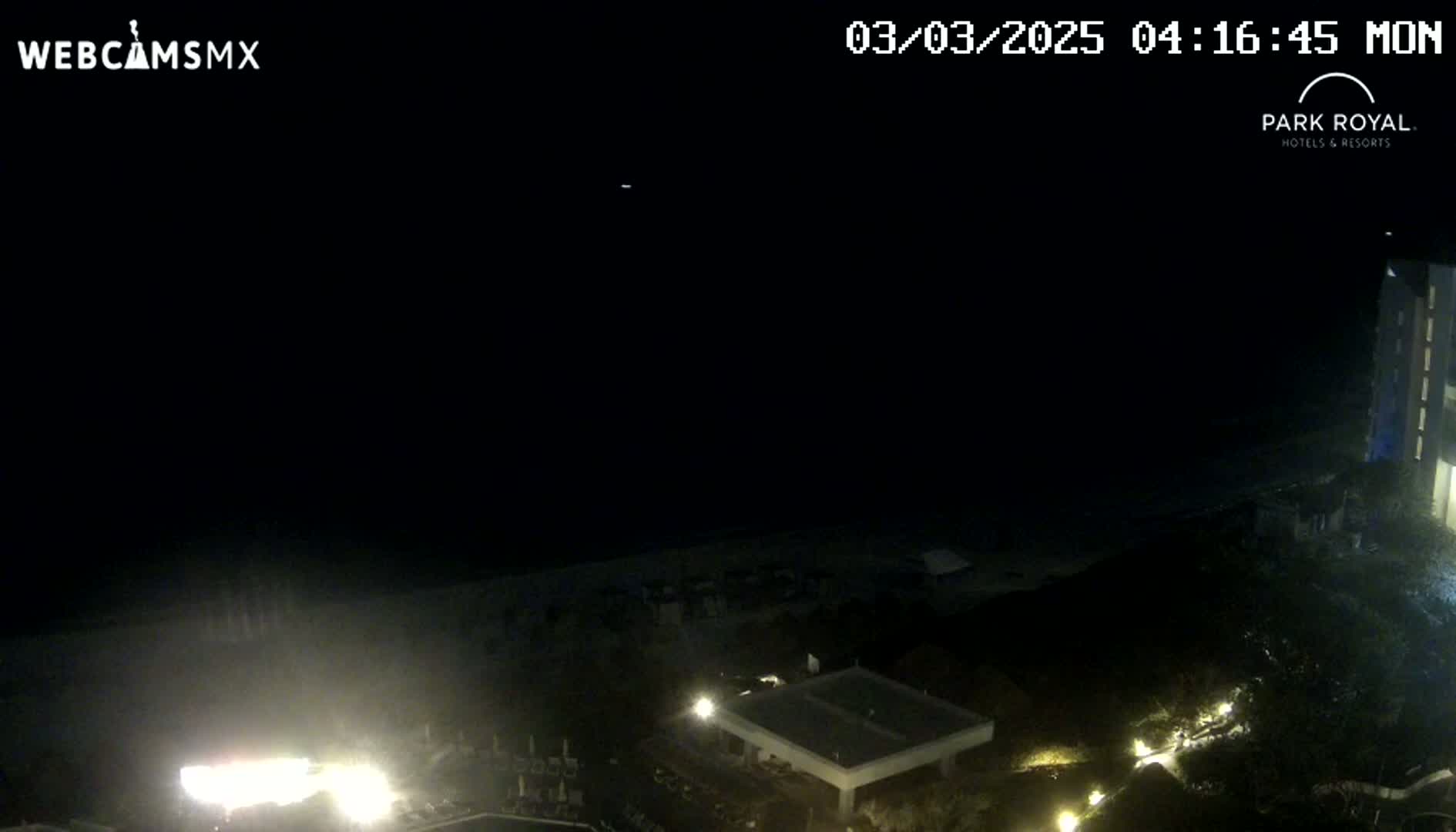 A nighttime, high-angle view shows a dark ocean, a beach, and dimly lit buildings and landscaping.