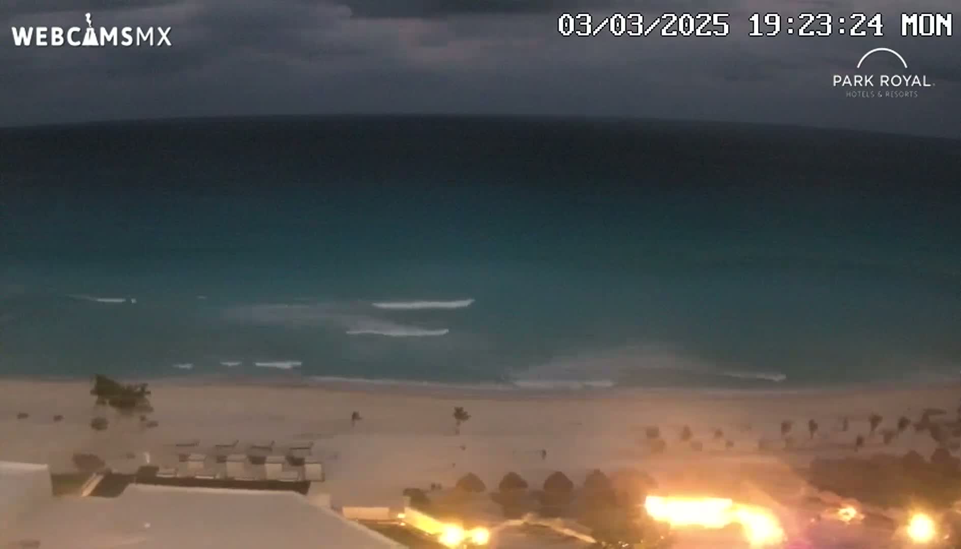 An elevated nighttime view shows a calm ocean meeting a sandy beach, with a resort and illuminated features in the foreground under a partly cloudy sky.