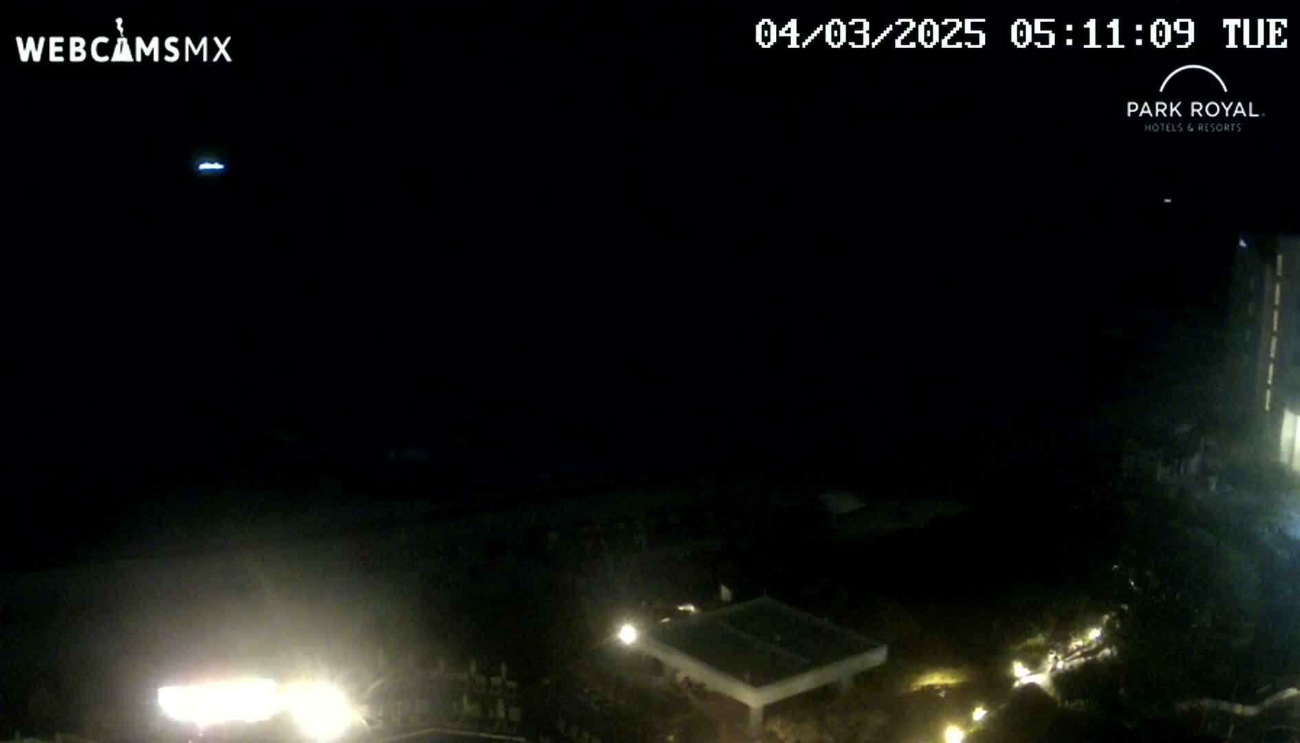 A nighttime, low-light view shows a dark ocean, sparsely lit grounds with some buildings, and a sliver of a tall building in the upper right corner.