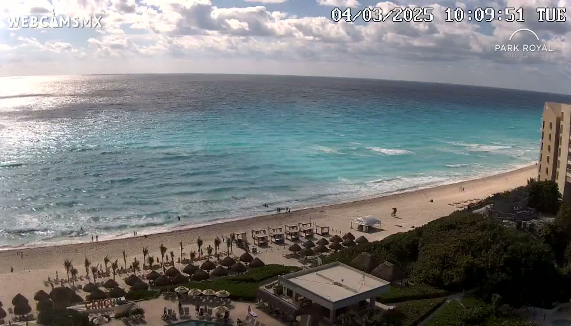 A partly cloudy day reveals a beach with turquoise water,  palapas, and lounge chairs, next to a resort building.