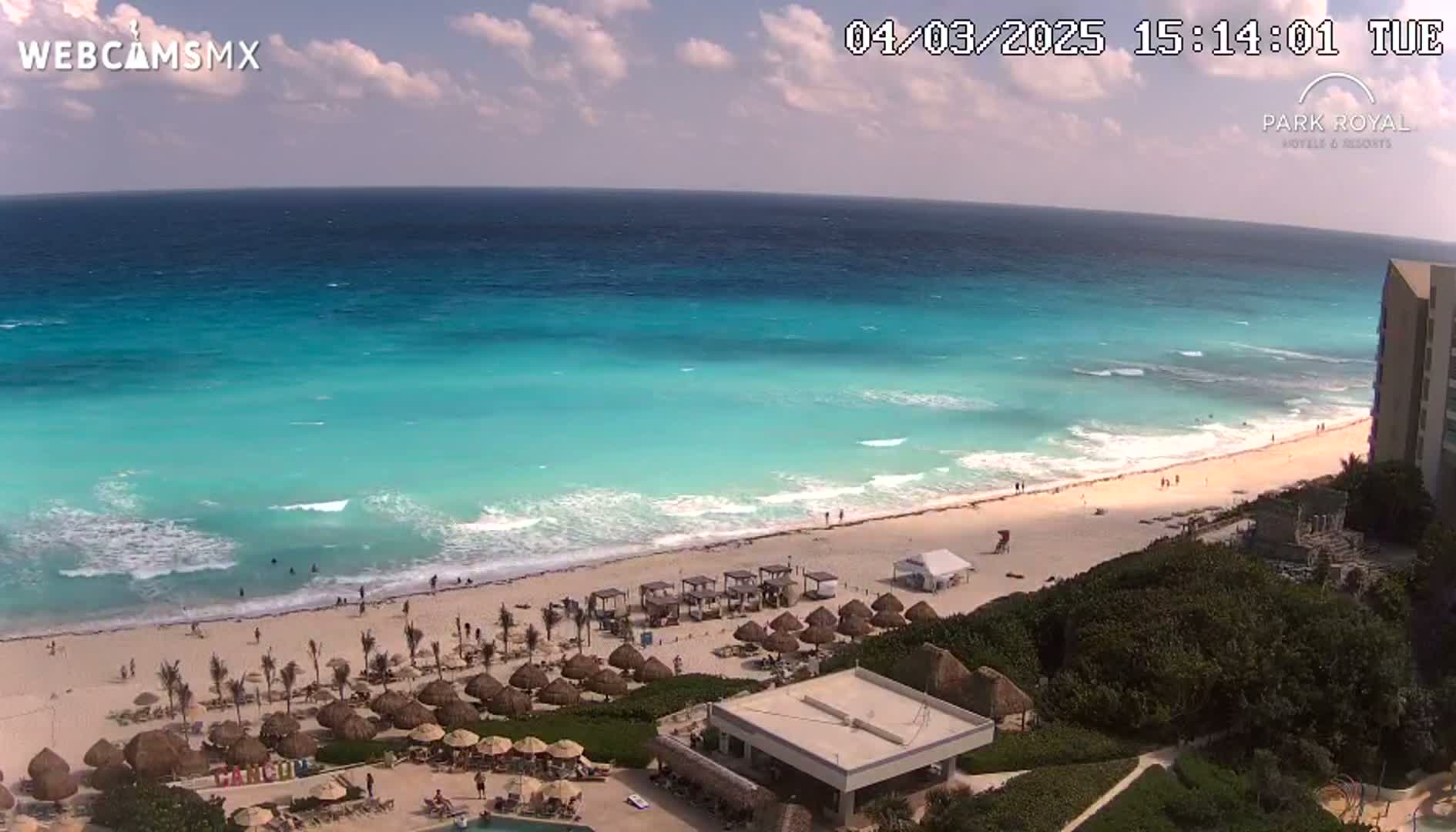 A partly sunny day shows a beach with turquoise water, small waves, and beach umbrellas in front of a resort building.