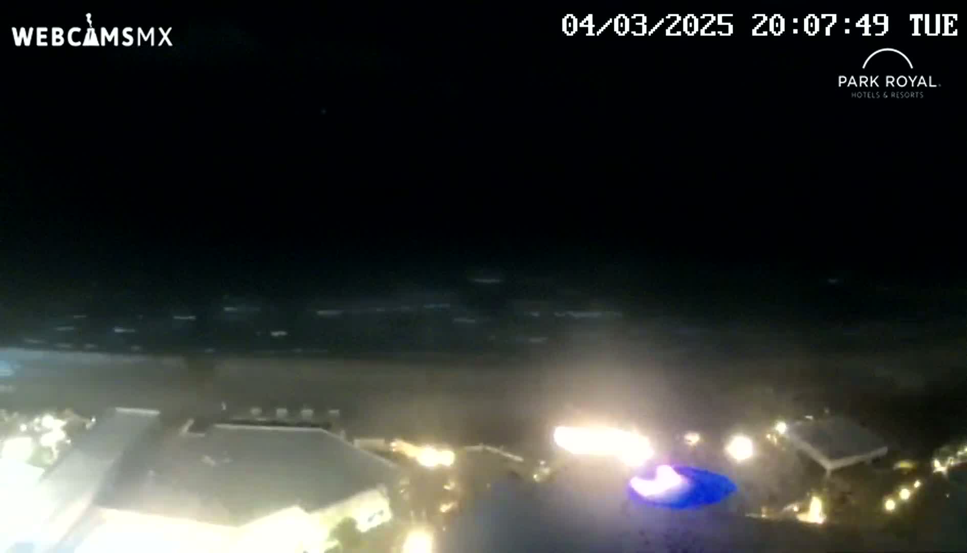A nighttime view of a beach with gently moving waves and illuminated buildings nearby.