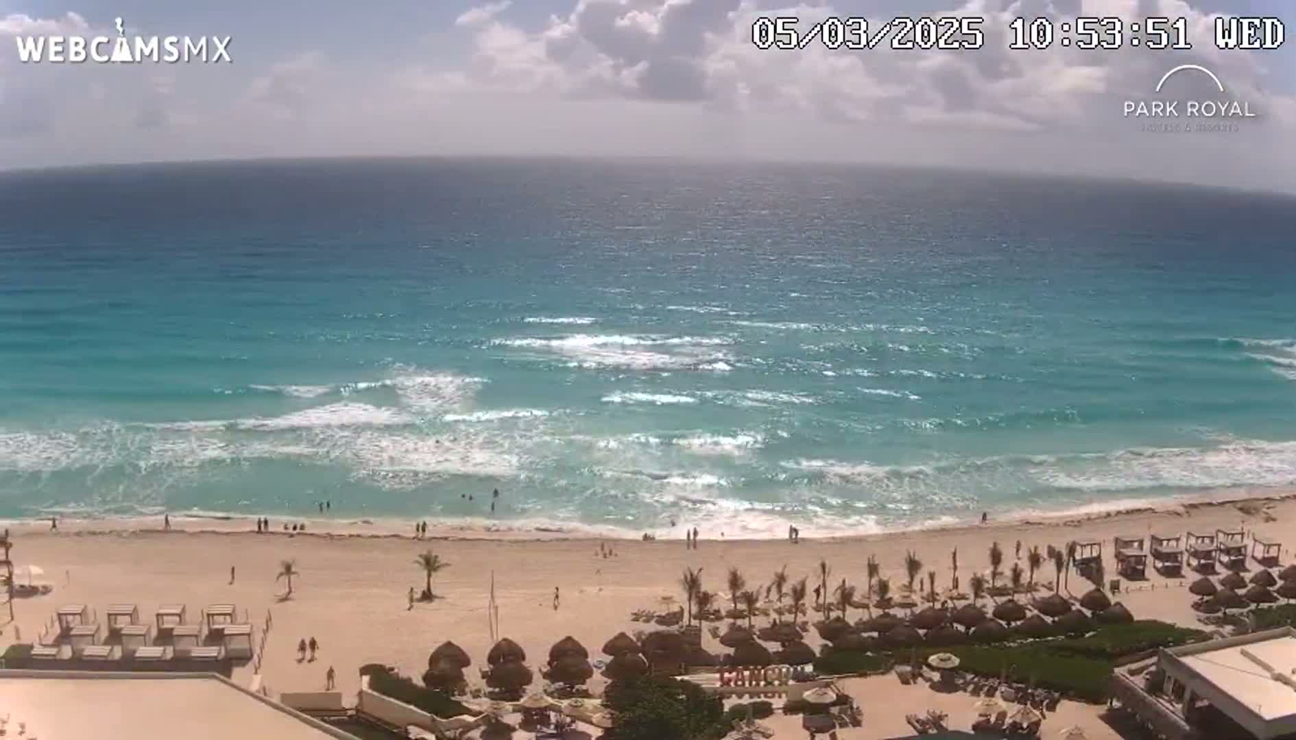 A beach with turquoise water and small waves under a partly cloudy sky is seen from an elevated vantage point, showing beach umbrellas and a resort in the foreground.