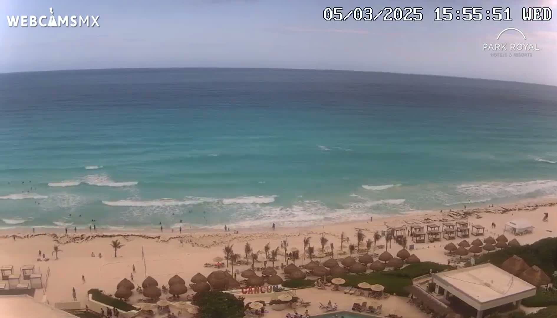 An aerial view shows a beach with turquoise water and small waves under a partly cloudy sky, with beach umbrellas and a resort visible.