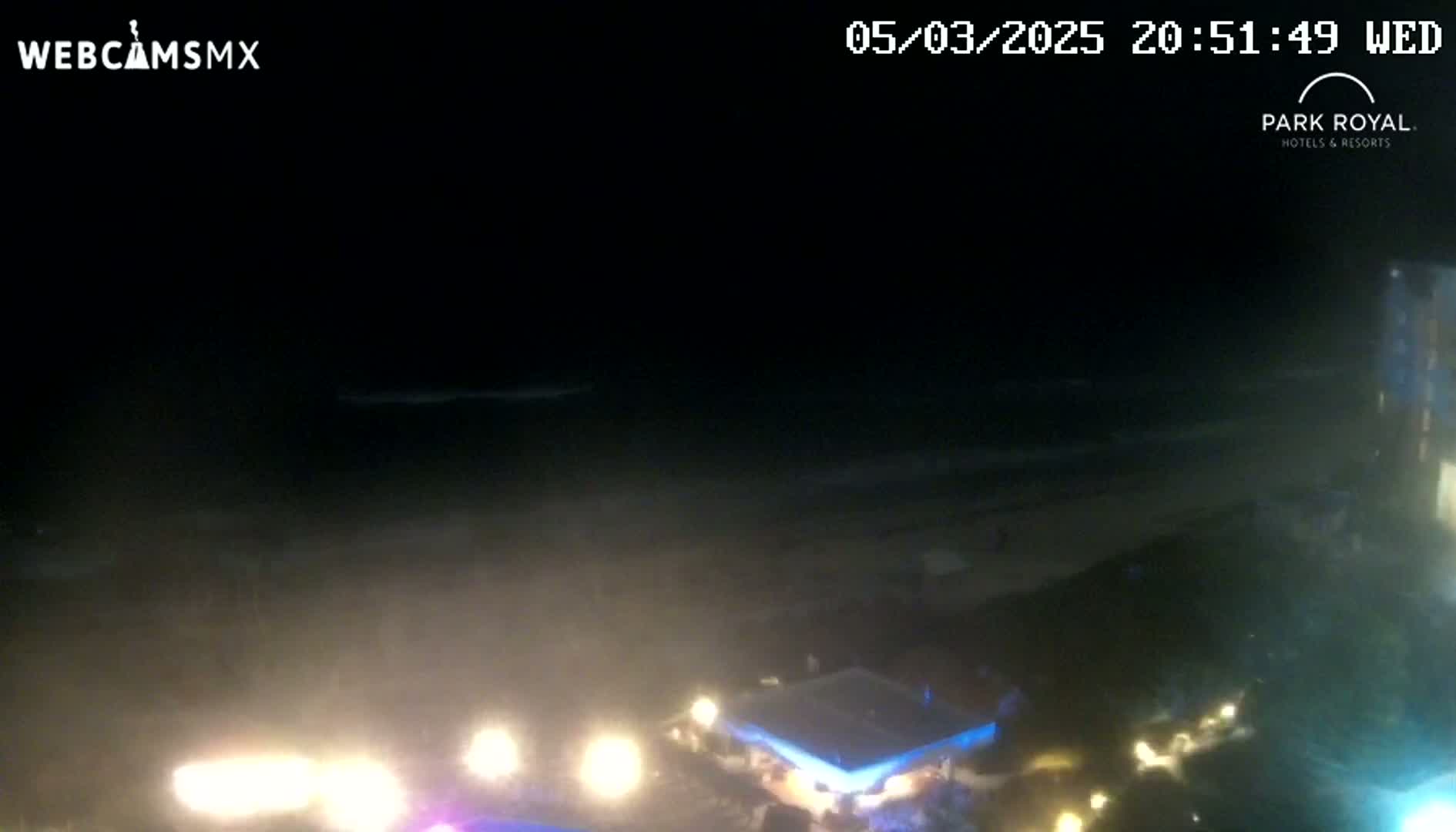 A nighttime view of a beach with dark, hazy weather conditions showing some lights from buildings and vehicles near the shore, and waves in the distance.