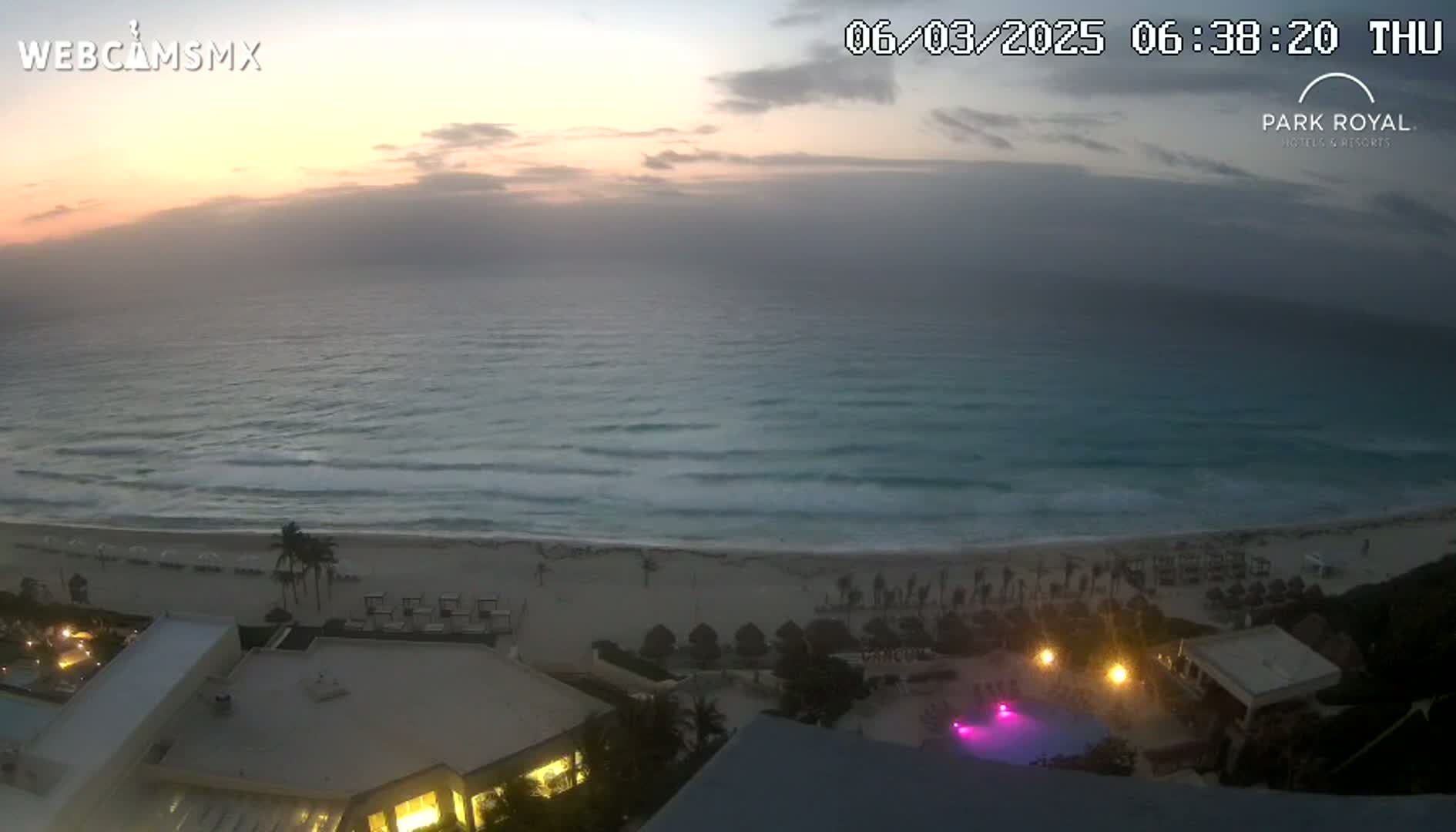 A hazy sunrise illuminates a beach with calm waves and a resort with lit pools.
