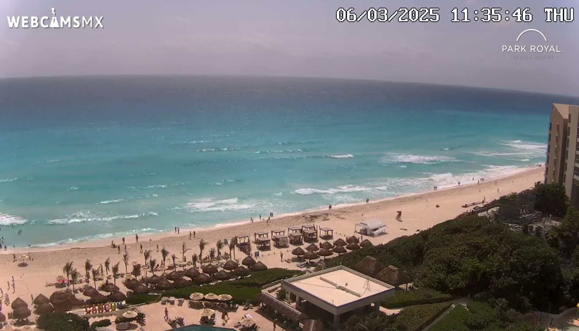 An aerial view shows a beach with turquoise water and small waves under a partly cloudy sky, with beach umbrellas and a resort visible.