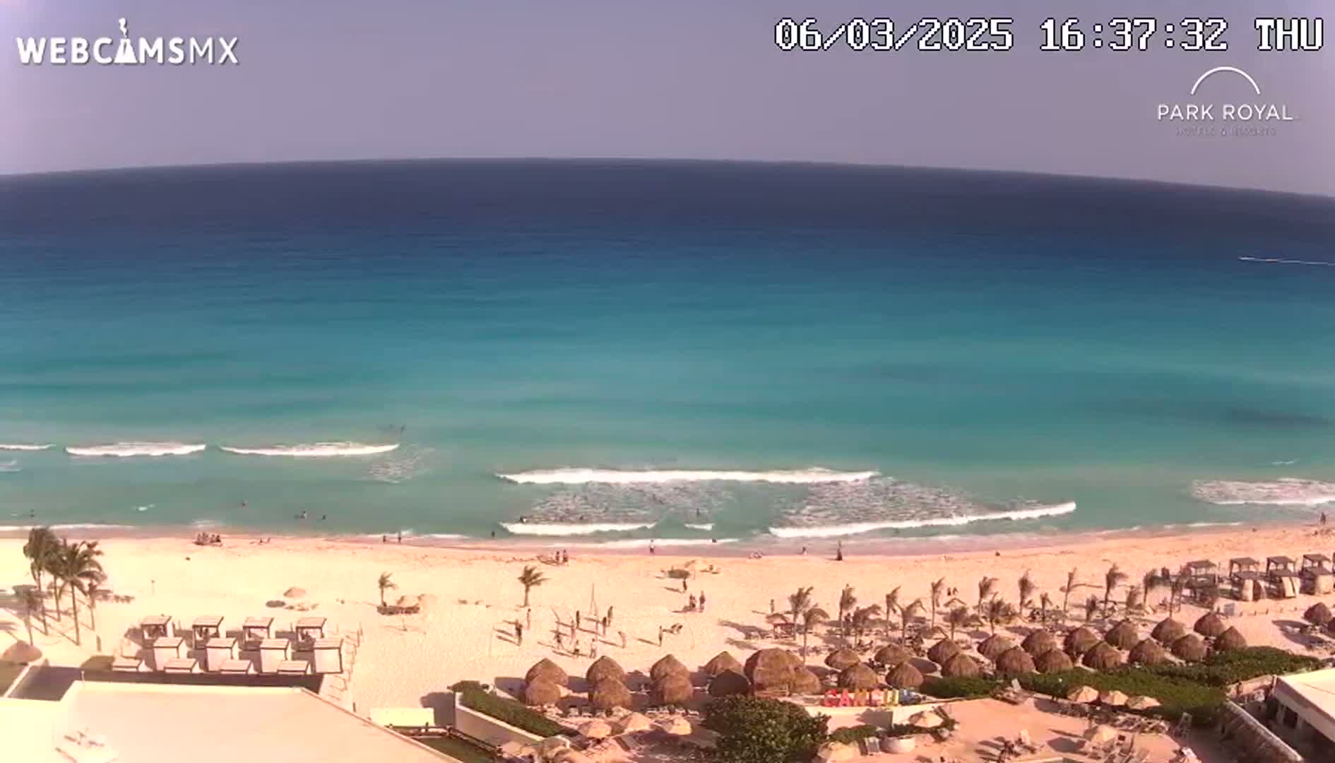 A mostly sunny day shows a clear turquoise ocean with small waves lapping a sandy beach lined with palm trees and thatched umbrellas.
