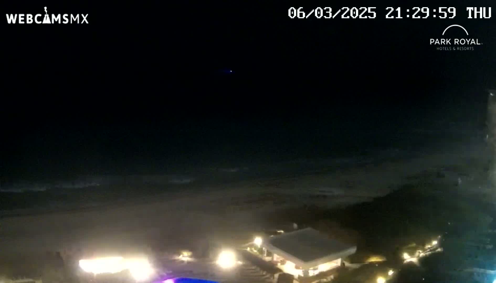 A nighttime view of a beach with gently lapping waves under a clear, dark sky shows illuminated buildings and landscaping near the shoreline.