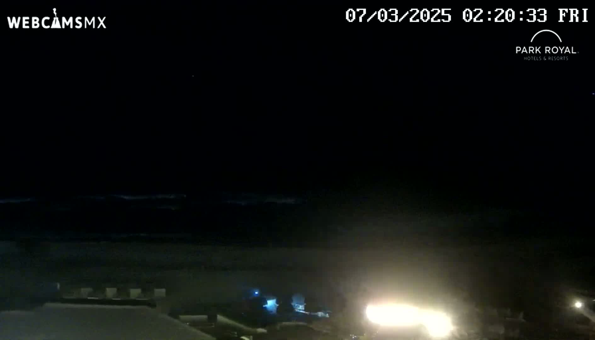 A nighttime coastal scene shows a dark ocean with waves and bright lights near the shoreline under a clear, dark sky.