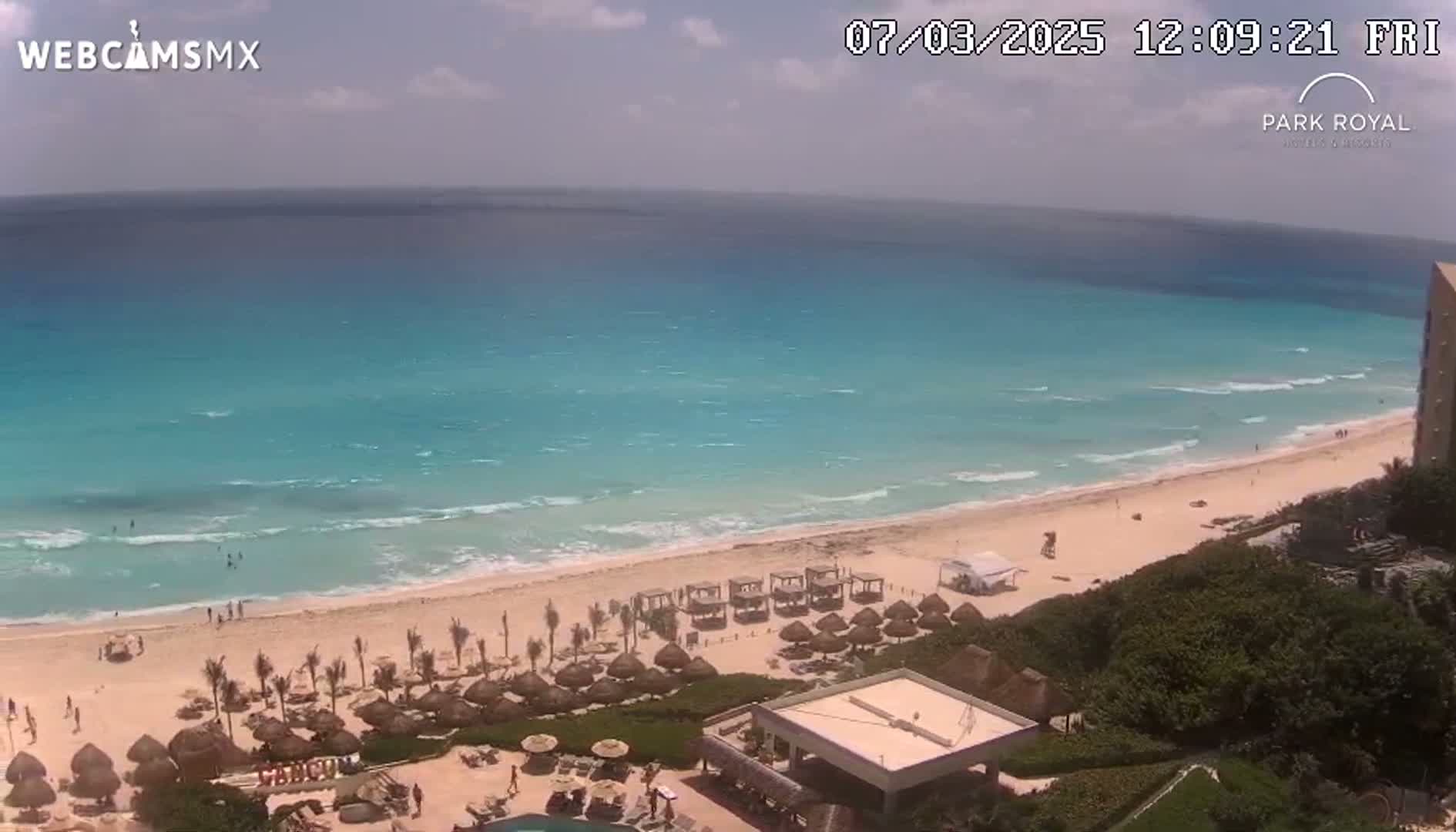 A partly sunny day shows a beach with calm waves, numerous beach umbrellas, and a resort.