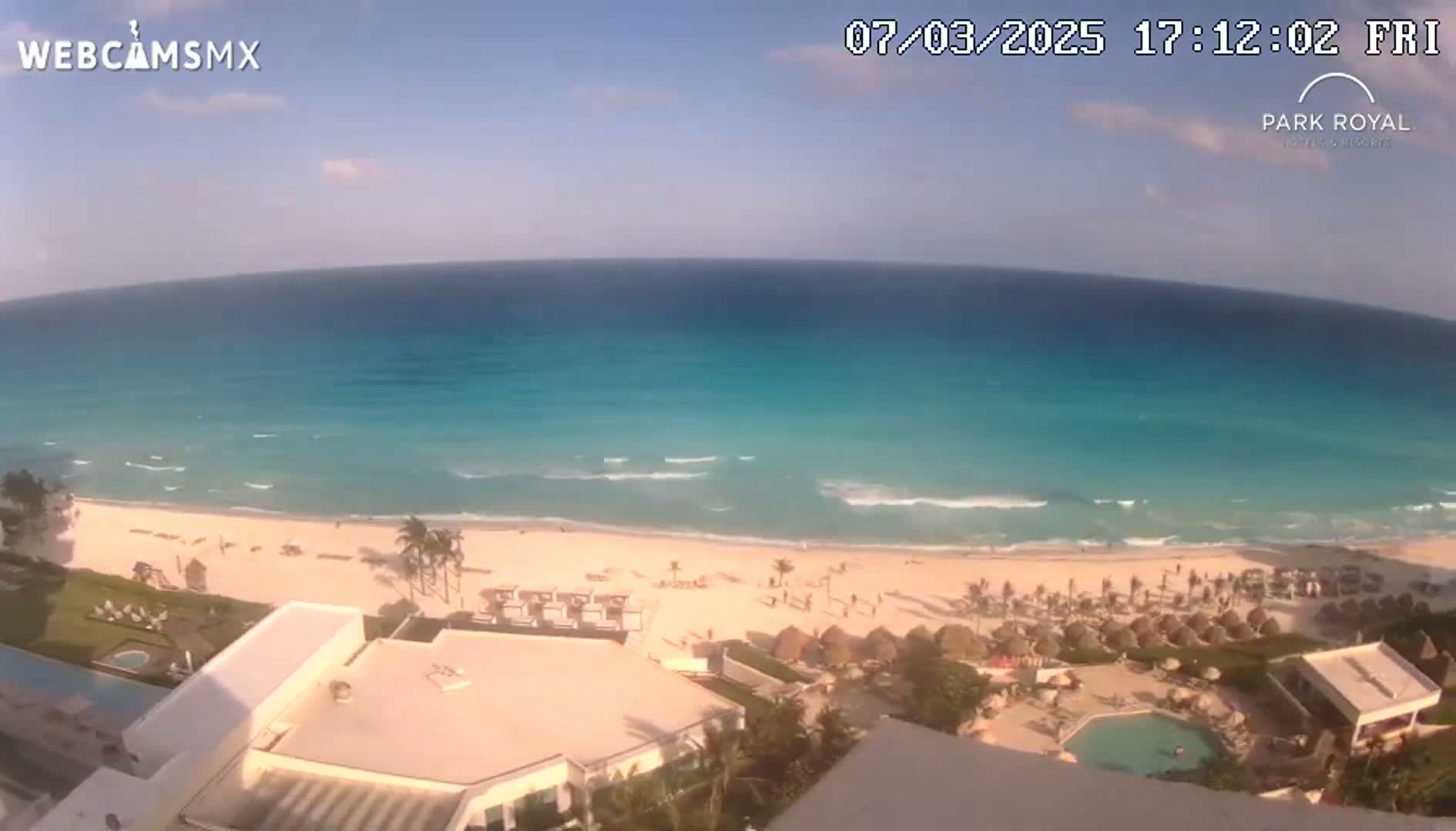 A mostly sunny day shows a beach with turquoise water, small waves, and beach umbrellas along a resort area.