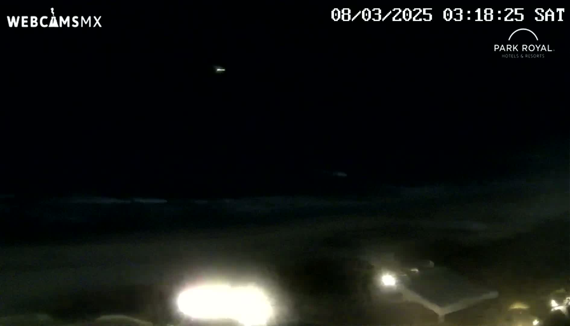 A nighttime, dark view of a beach with faint waves and a few distant lights visible.