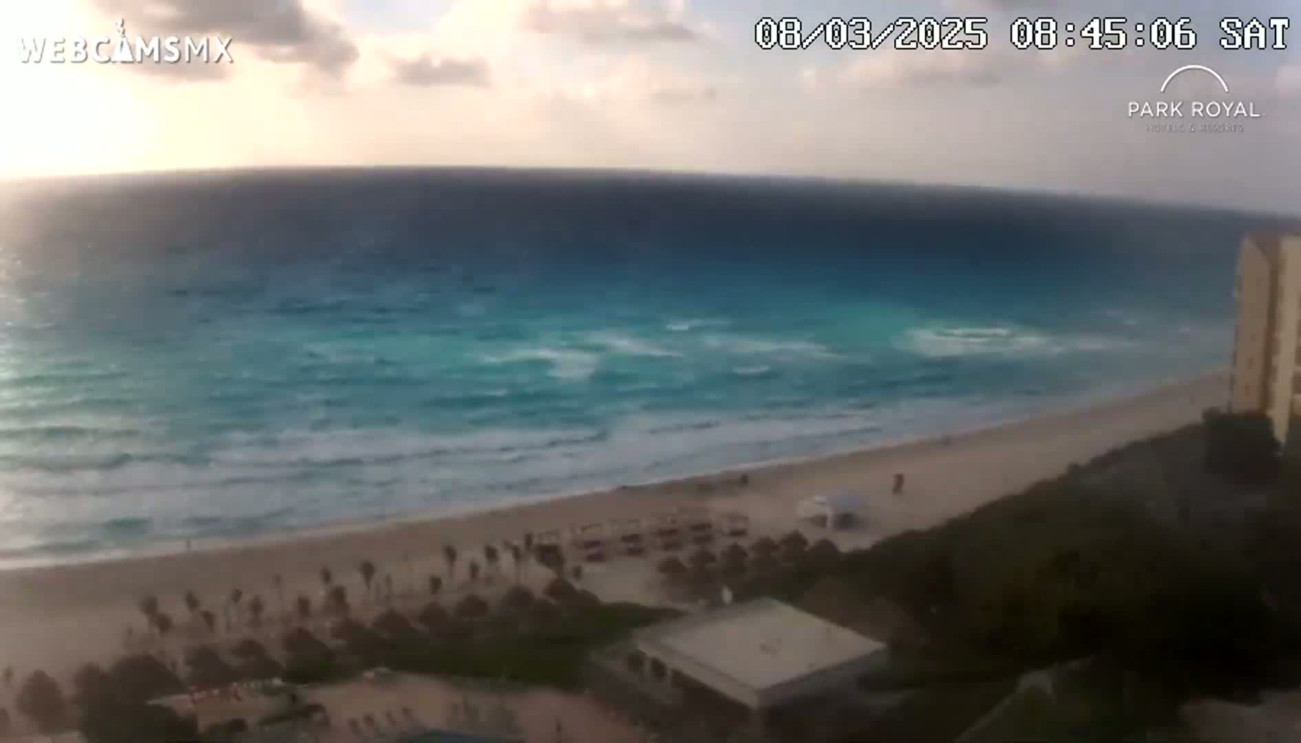 A mostly clear sky overlooks a beach with calm waves and tropical vegetation under partly cloudy conditions.