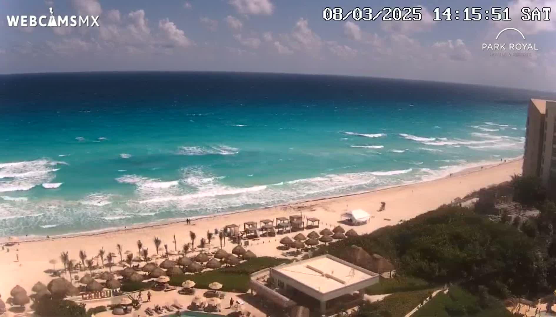 An aerial view shows a beach with turquoise water and moderate waves under a partly cloudy sky.