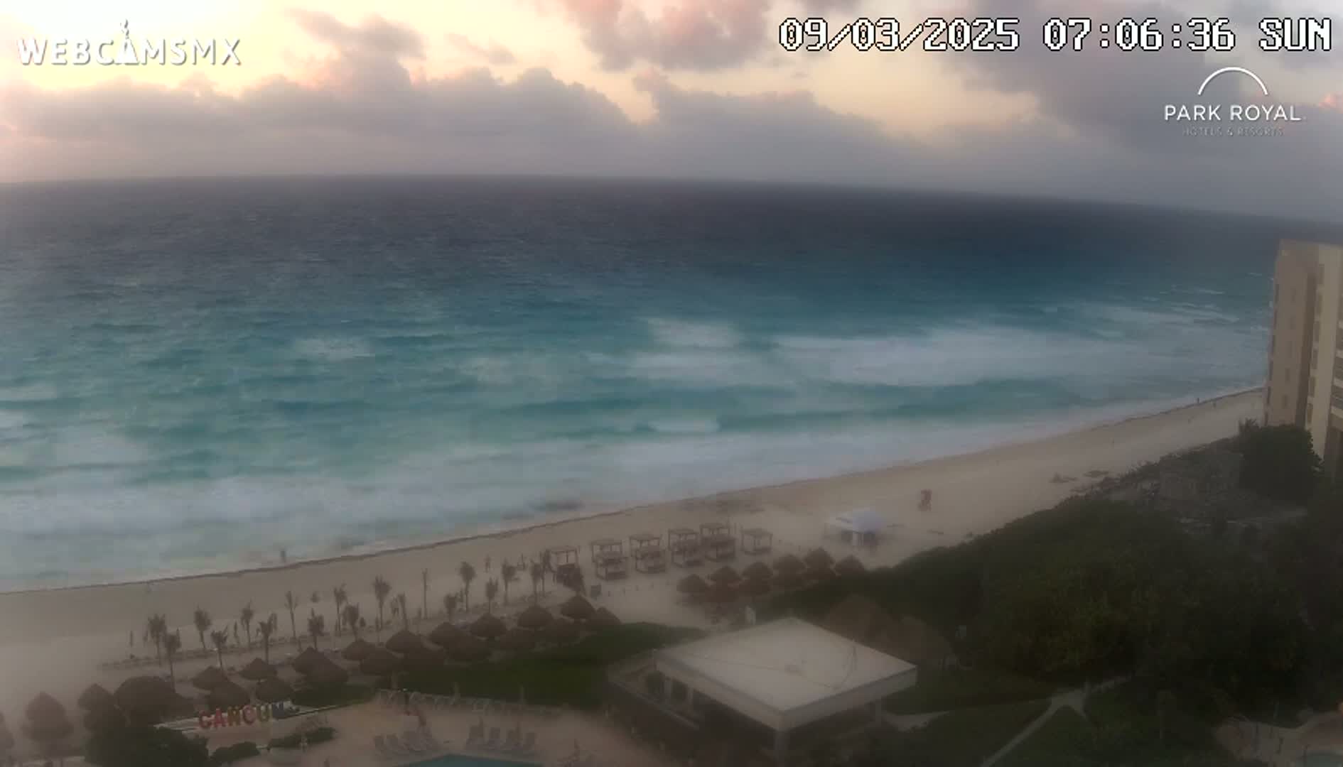 A beach with turquoise water and moderate waves under an overcast sky is seen from a high vantage point, showing beach umbrellas, buildings, and vegetation.