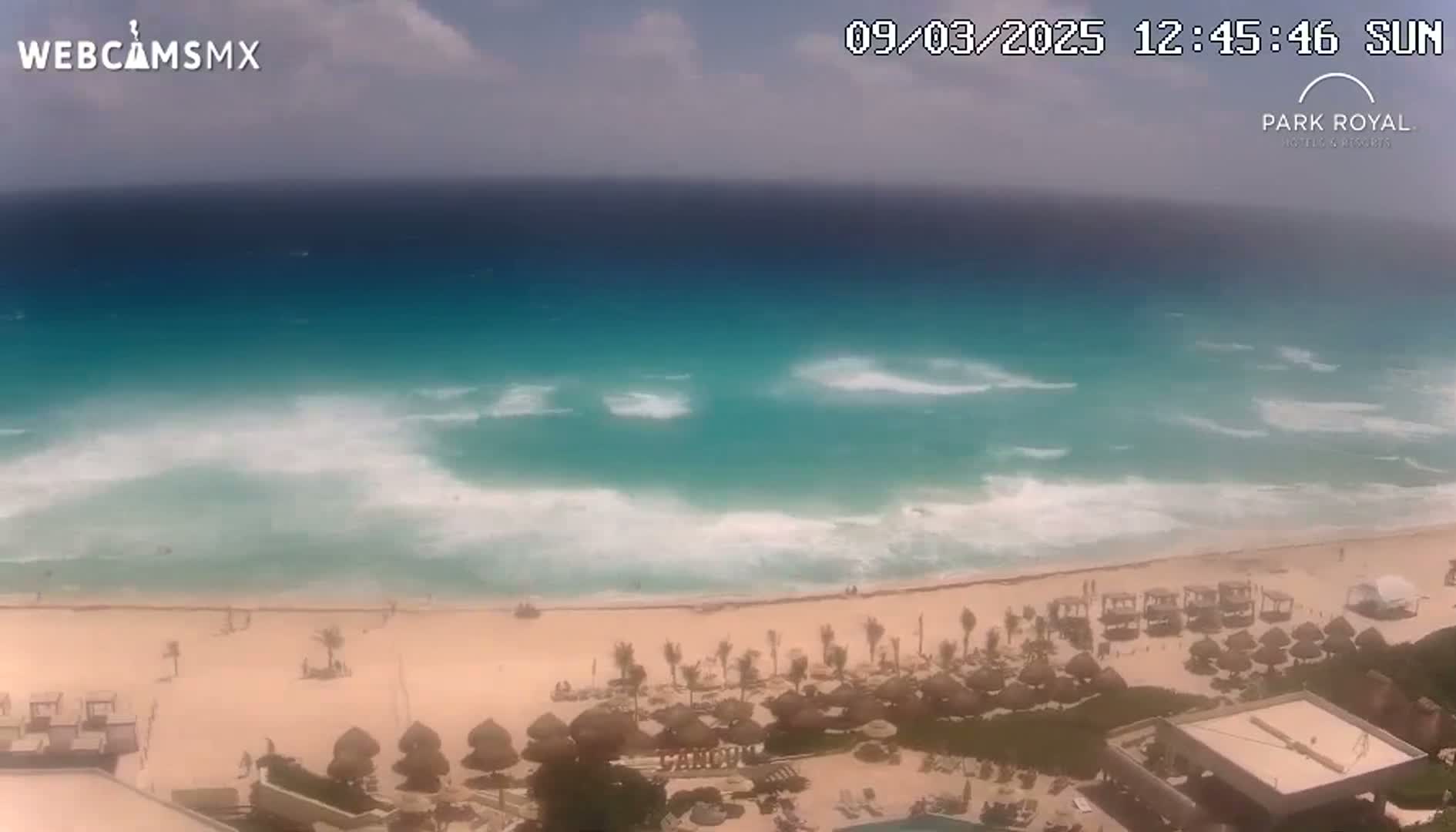 A beach with turquoise water and moderate waves under partly cloudy skies is seen from an elevated viewpoint, showing beach umbrellas and resort buildings.