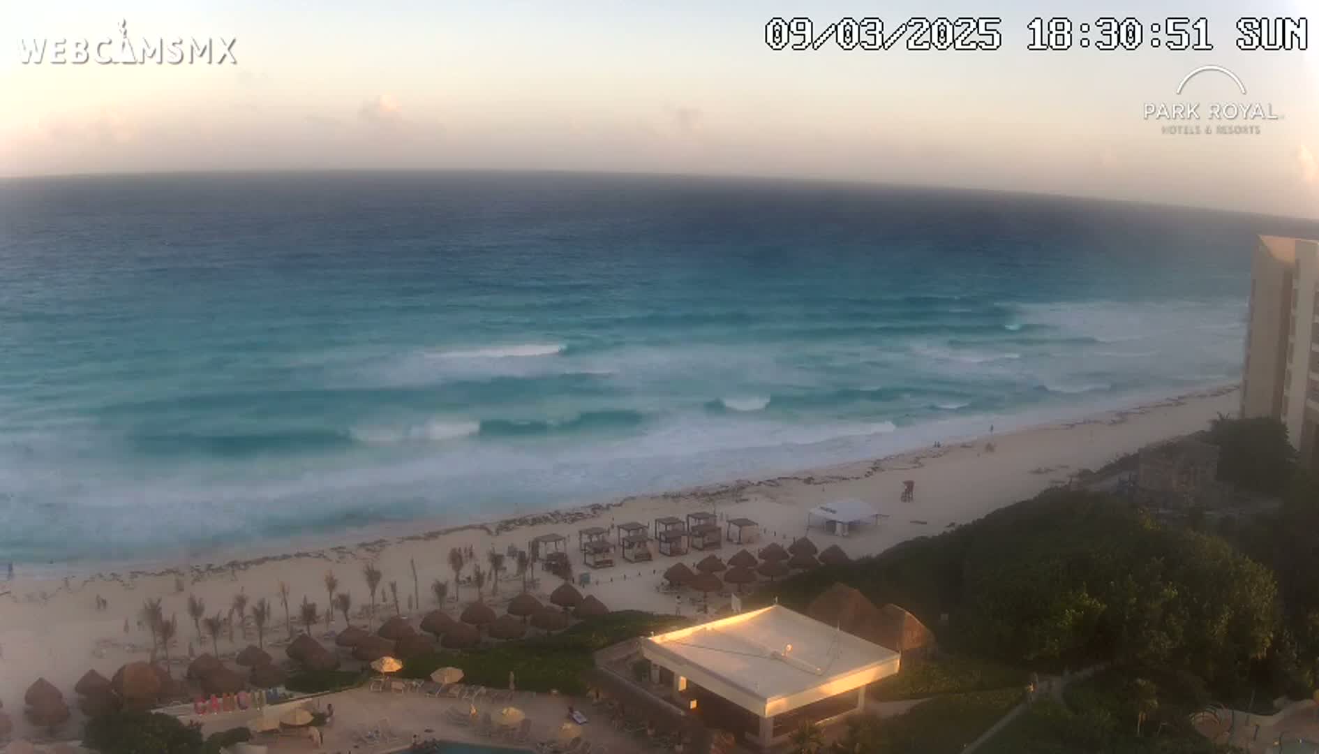 A beach with turquoise water and moderate waves is seen at sunset, with beach umbrellas and a building visible in the foreground.