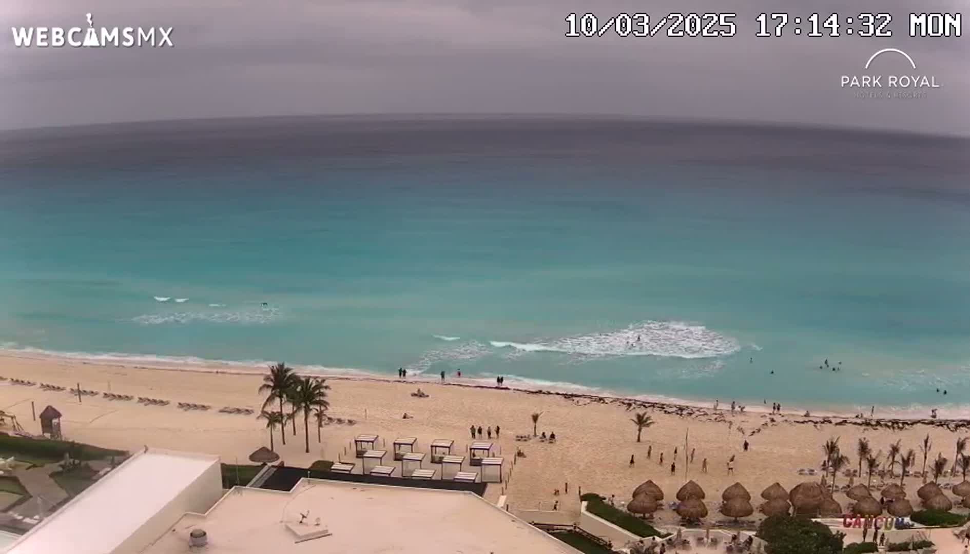 An aerial view shows a beach with turquoise water under an overcast sky, palm trees, and beach umbrellas.