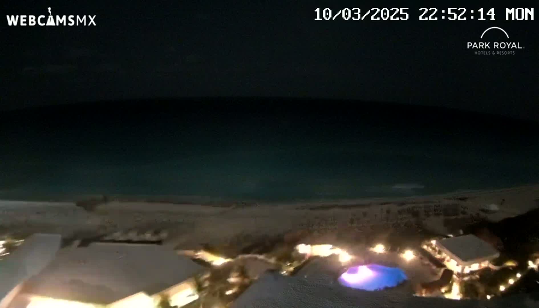 A nighttime, high-angle view shows a dark ocean meeting a light-colored beach, with illuminated buildings and a pool visible in the foreground.