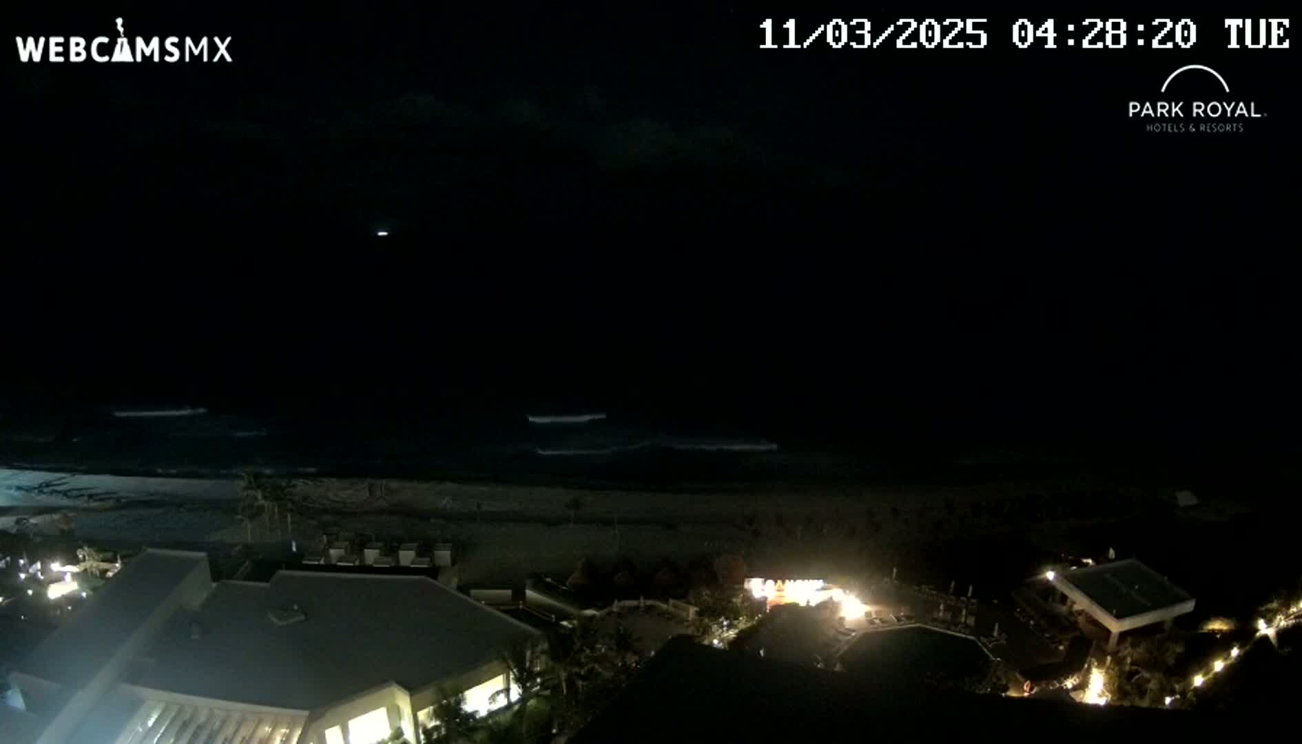 A nighttime, overhead view shows a dark ocean with small waves lapping a beach, and a resort with illuminated buildings and landscaping.