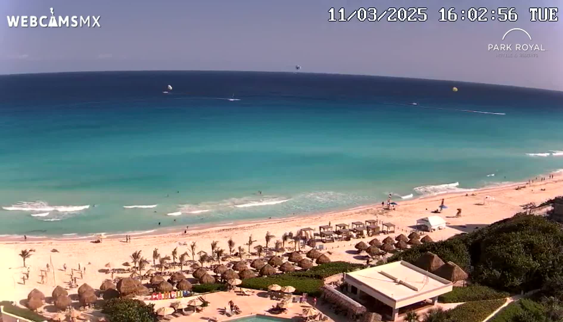 A clear, sunny day shows a beach with turquoise water, gentle waves, and thatched umbrellas lining the shore, adjacent to a resort with a pool.