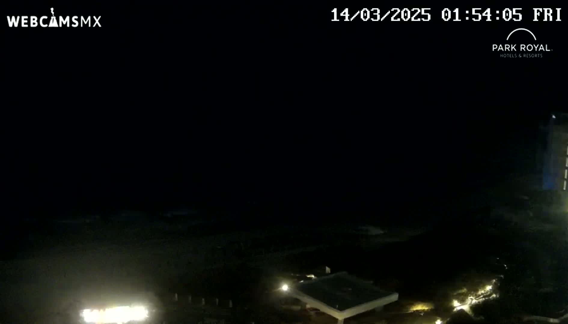 A nighttime, low-light view of a dark beach and ocean with some lights visible from buildings and possibly vehicles nearby.