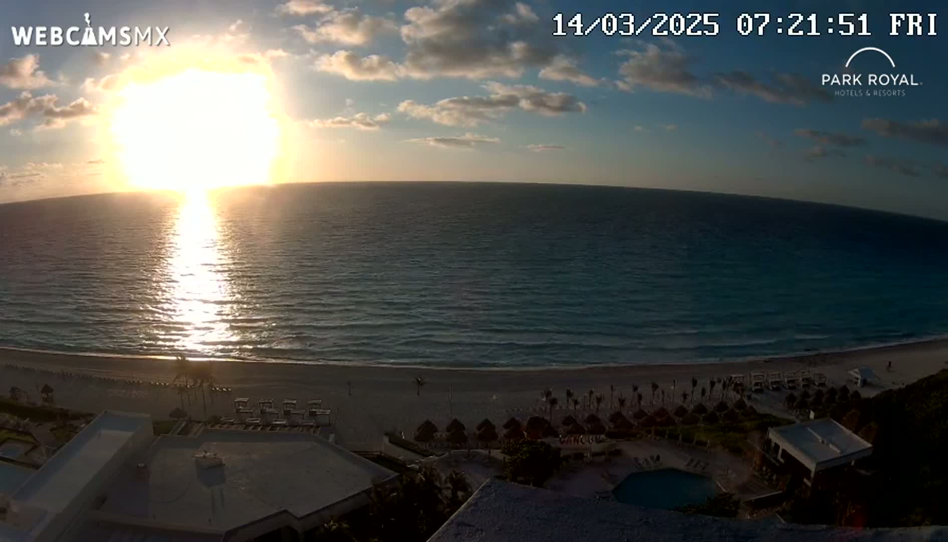 A sunrise over a calm ocean, illuminating a beach with resort buildings and a swimming pool.